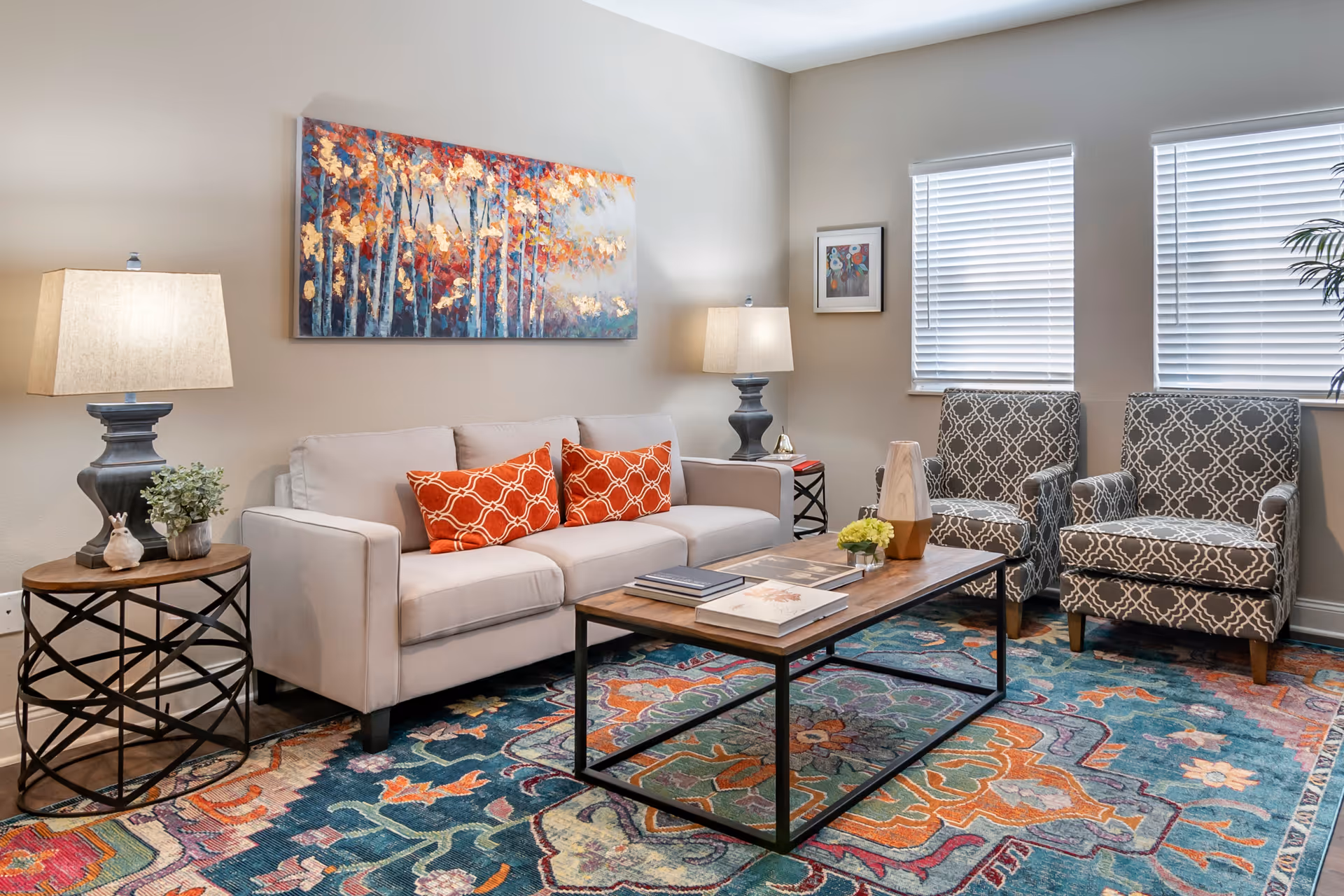 A cozy living room with a beige sofa adorned with two orange patterned pillows, two gray patterned armchairs, a wooden coffee table with books and decorative items, two side tables each with a lamp, a colorful patterned rug, and wall art including a large painting of autumn trees and a smaller framed picture near two windows with white blinds.