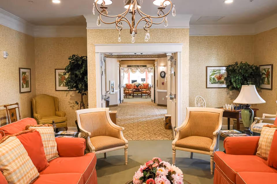 A cozy senior living facility common area with two orange sofas and two beige armchairs arranged around a coffee table with a floral centerpiece. The room has warm yellow wallpaper, framed botanical prints on the walls, and a chandelier hanging from the ceiling. In the background, a hallway leads to another seating area with more chairs and a window letting in natural light.