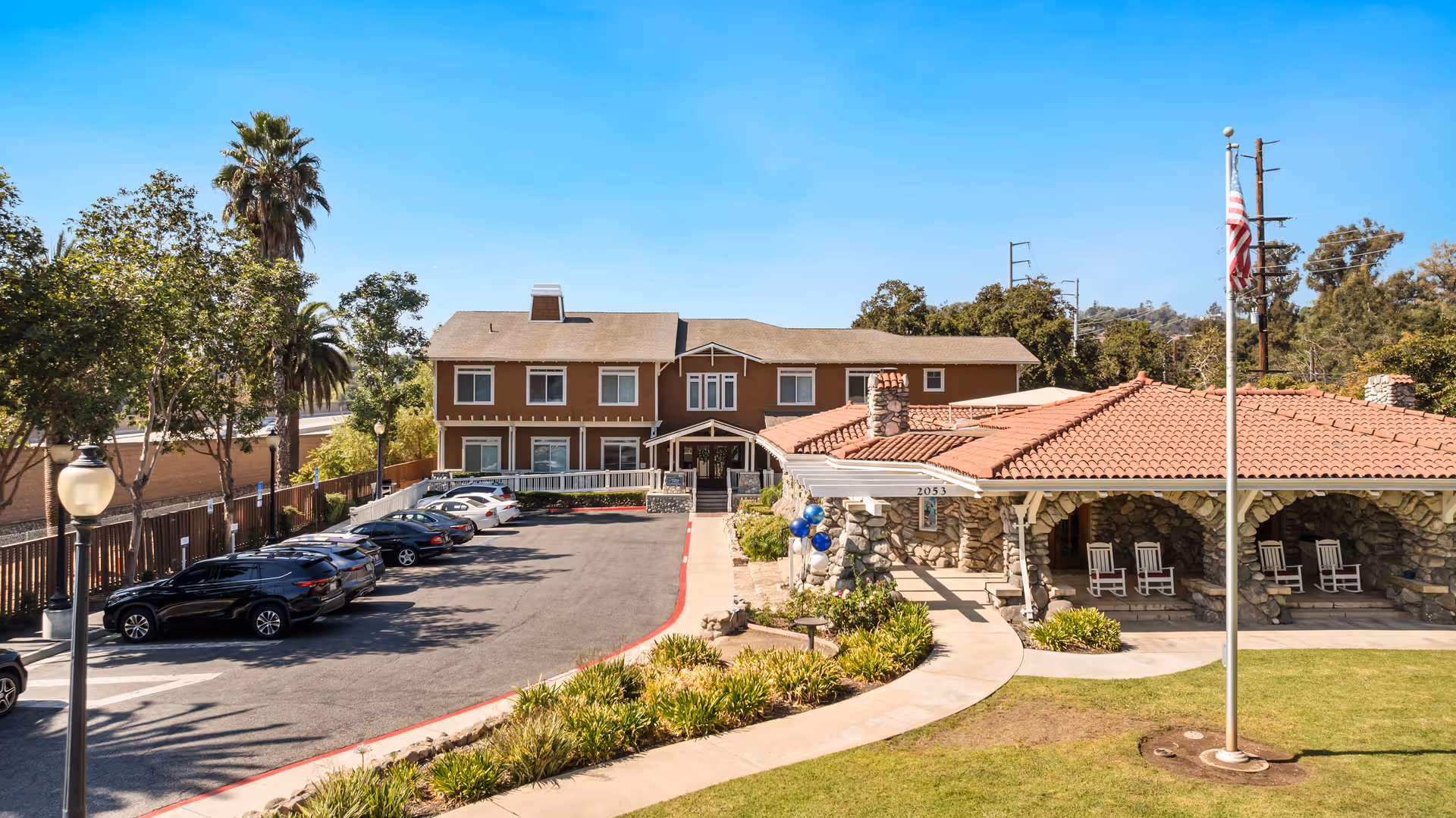 Exterior view of Ivy Park at Claremont senior living facility showing a two-story brown building with a parking lot on the left and a stone building with a red tiled roof and rocking chairs on the right. There is a flagpole with an American flag in front of the stone building, surrounded by green grass and landscaping under a clear blue sky.