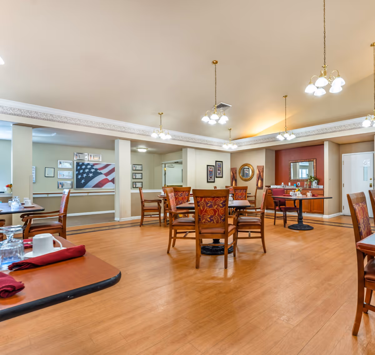 A spacious dining room with wooden floors and several tables and chairs arranged for meals. The chairs have patterned upholstery. The room is well-lit with multiple hanging light fixtures. There is a sideboard with a mirror and decorative items against one wall, and framed pictures and an American flag artwork are displayed on the walls.