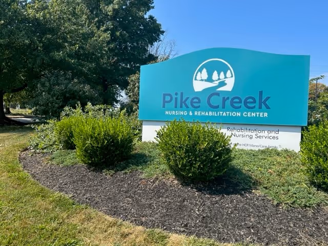 Outdoor view of a sign for Pike Creek Nursing & Rehabilitation Center surrounded by green bushes and trees under a clear blue sky.