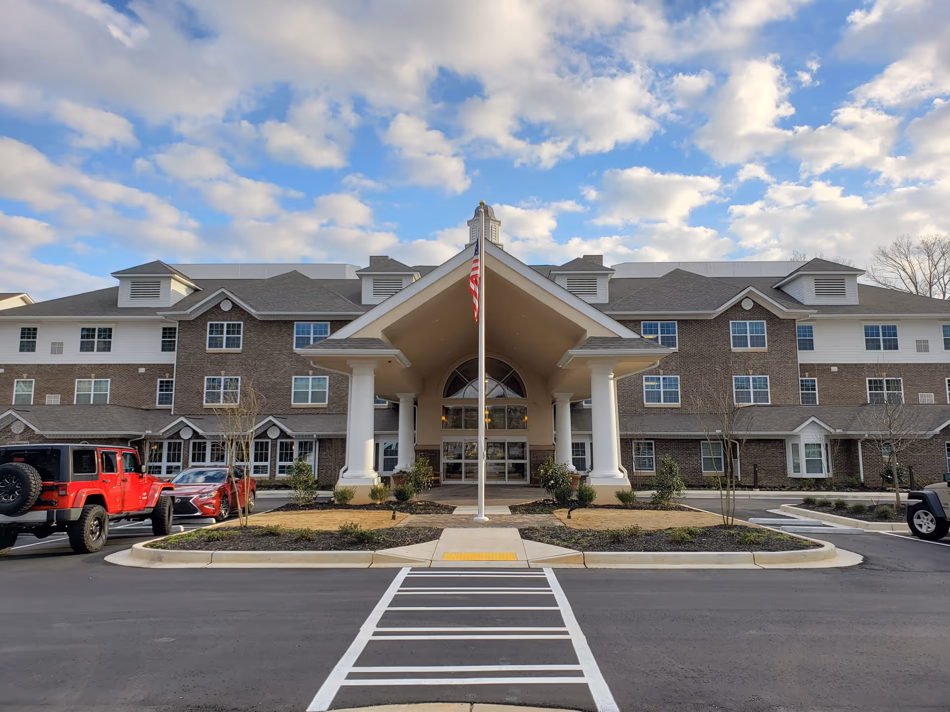 Front exterior view of Vickery Rose Retirement Resort building with a covered entrance supported by white columns, an American flag on a flagpole in front, several parked cars, and a partly cloudy sky above.