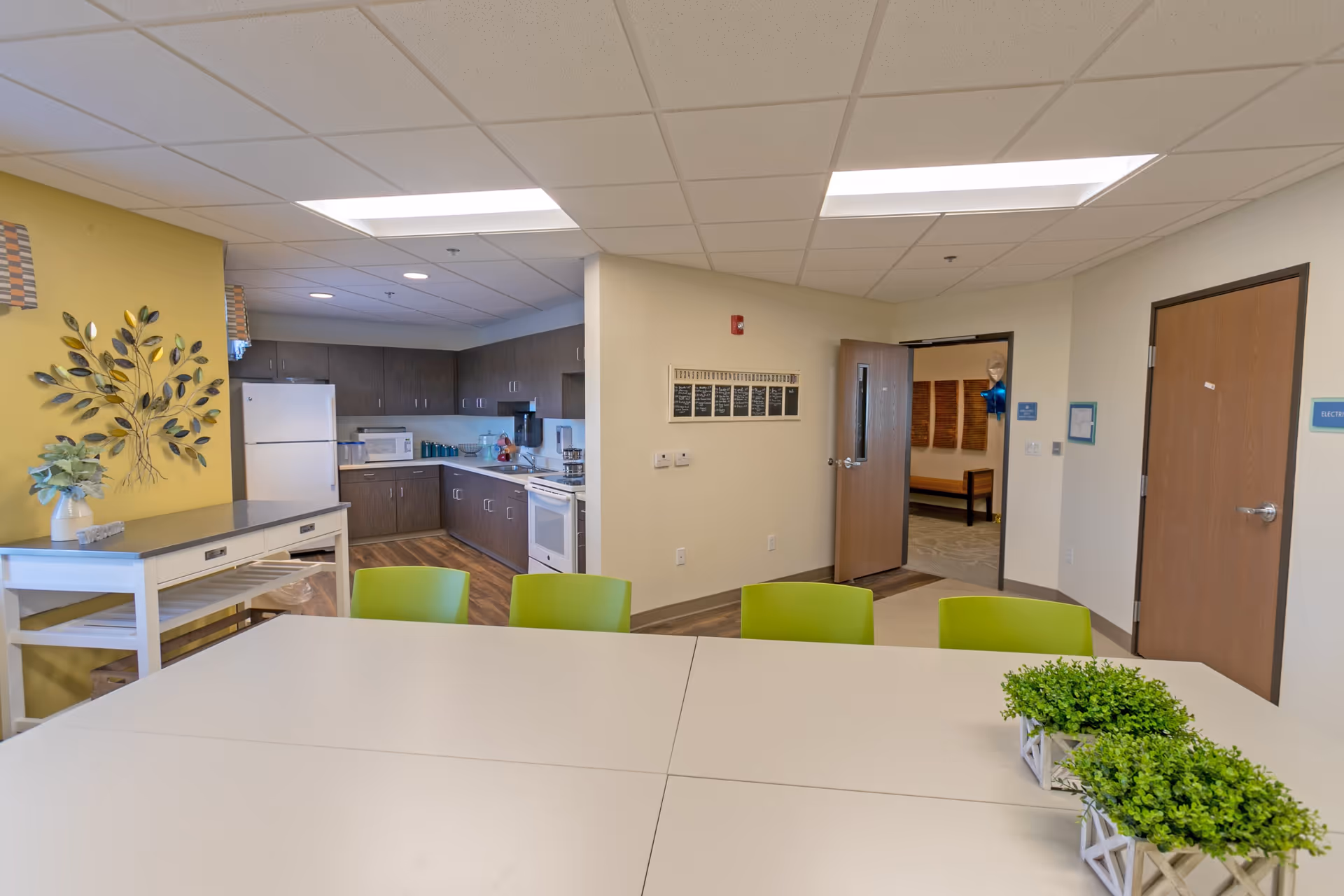 Interior view of a senior living facility kitchen and dining area with a white table surrounded by green chairs, a kitchen with dark cabinets, white appliances, and a yellow accent wall with decorative metal leaves and a plant on a small table.