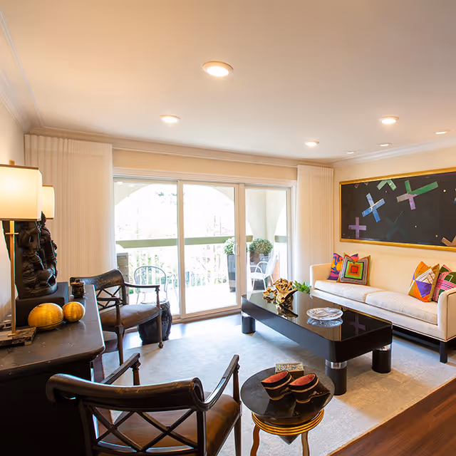 Bright living room with a sofa and colorful pillows, a black coffee table, chairs, and sliding glass doors opening to a balcony.