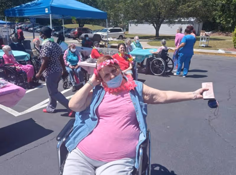 A group of elderly people, some in wheelchairs, gathered outdoors in a parking lot area with tables covered in colorful tablecloths. Several caregivers are present, and one elderly woman in the foreground is wearing a pink shirt, a flower headband, a face mask, and a lei, smiling and holding out a phone case. There is a blue canopy tent providing shade over some of the tables, and cars and trees are visible in the background.