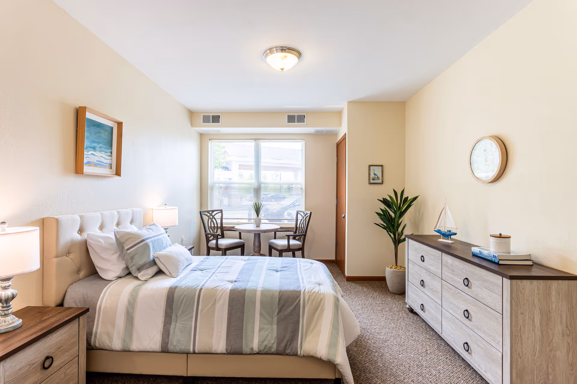 A bright and neatly arranged bedroom in a senior living facility featuring a bed with striped bedding, two bedside tables with lamps, a small round table with two chairs near a window, a dresser with decorative items including a small sailboat model, and a potted plant in the corner.