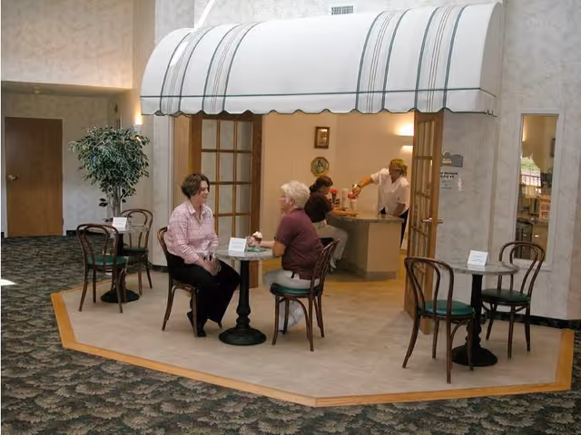 A small café-style seating area inside a senior living facility with three women sitting at two tables, one woman behind a counter serving drinks, and a white awning overhead. The area has wooden chairs with green cushions and a potted plant nearby.
