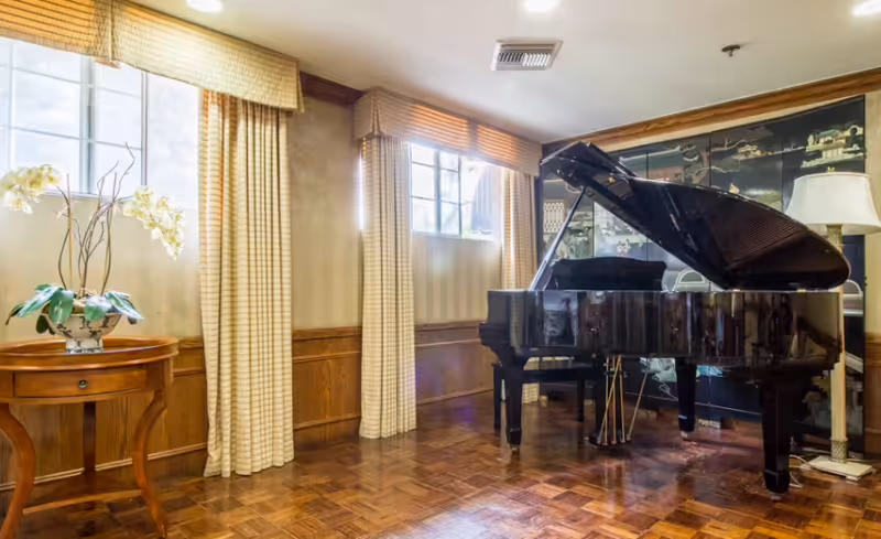 A bright room with large windows covered by beige curtains, featuring a black grand piano with its lid open, a wooden side table with a white orchid plant, and a floor lamp next to a decorative black folding screen with gold designs.