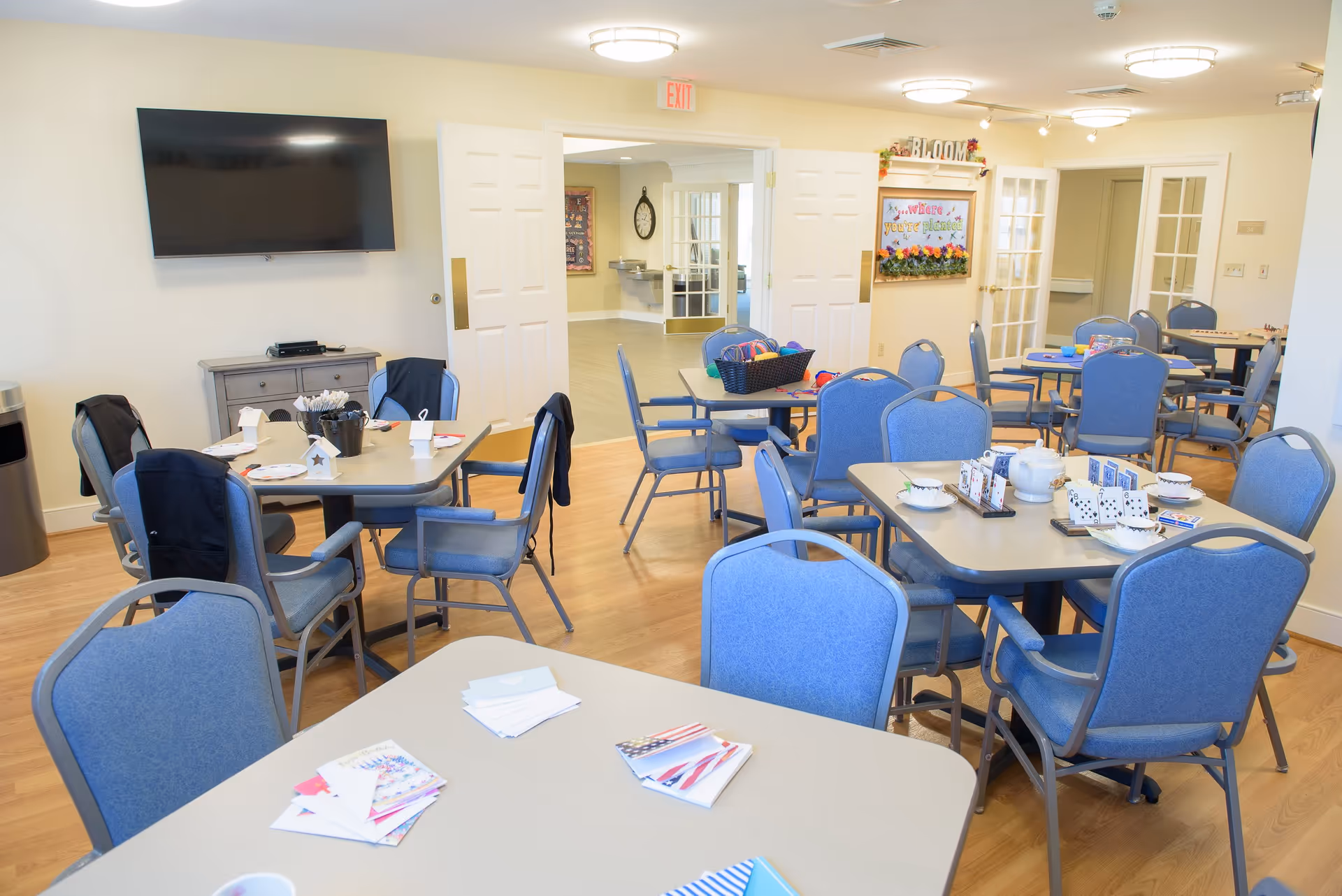 A bright, spacious common room with multiple tables and blue cushioned chairs arranged for group activities. Tables have various items such as playing cards, tea sets, craft supplies, and booklets. A large flat-screen TV is mounted on the wall above a small cabinet. Double doors lead to another room with water fountains and a clock on the wall. A decorative sign on the wall reads 'BLOOM ...where you're planted.'