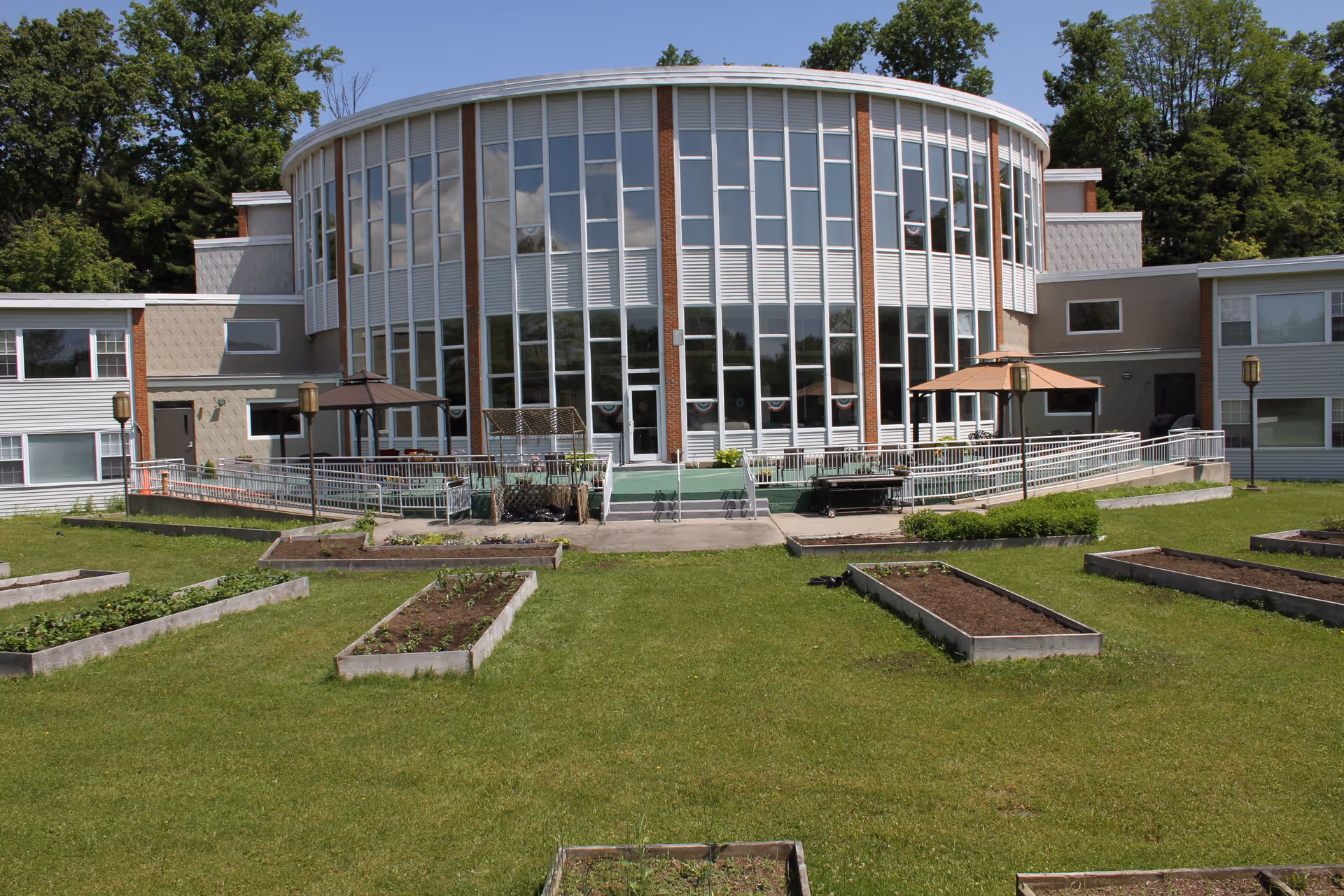 Exterior view of a senior living facility with a large curved glass window section in the center, surrounded by two wings of the building. In front of the building, there is a well-maintained lawn with multiple raised garden beds and two patio umbrellas providing shade over seating areas. Trees and greenery are visible in the background.