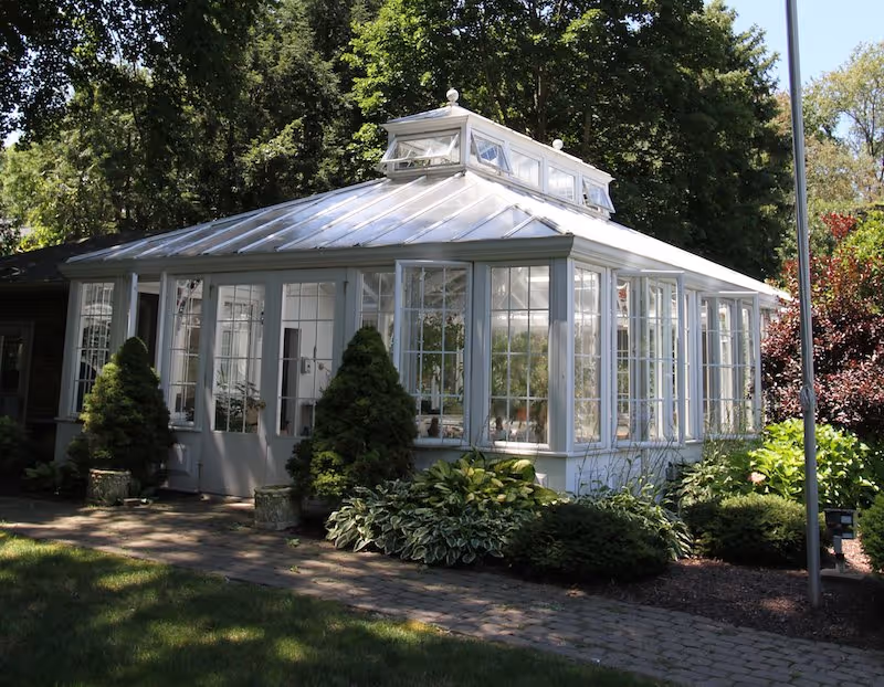 A white greenhouse-style building with large windows and a glass roof, surrounded by green shrubs and trees on a sunny day.