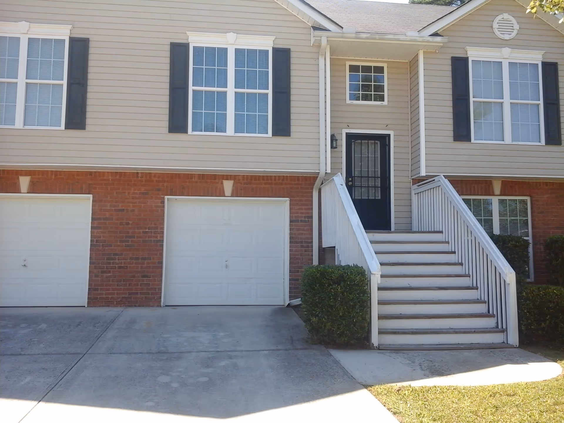 Exterior view of a residential building with beige siding and red brick lower walls. The building has two white garage doors, a black front door accessed by a white staircase with railings, and several windows with white frames and black shutters. There are small bushes near the stairs and driveway.