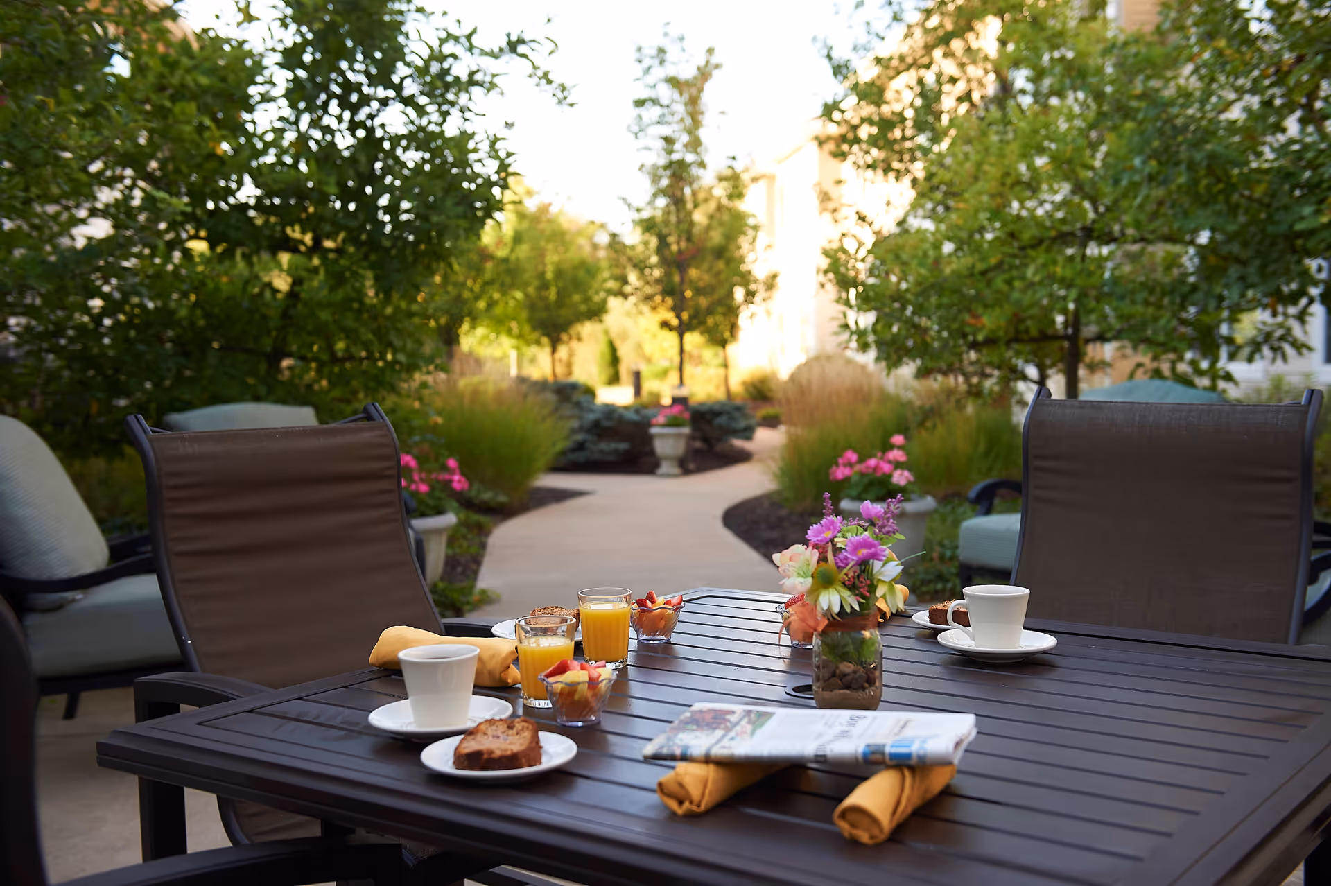 Outdoor patio area with a dark wooden table set with two cups of coffee, two glasses of orange juice, small bowls of fruit, a vase with colorful flowers, and a folded newspaper. The table is surrounded by cushioned chairs and the background shows a garden path lined with trees and plants.