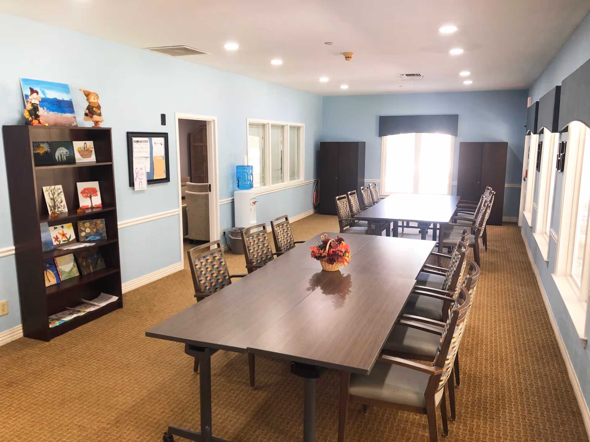 A well-lit room with two long tables arranged in a row, surrounded by chairs. A small basket with artificial flowers is placed on the nearest table. The room has light blue walls, a brown carpet, and a bookshelf displaying various paintings and crafts. There is a water dispenser against the wall and two large windows at the far end letting in natural light.