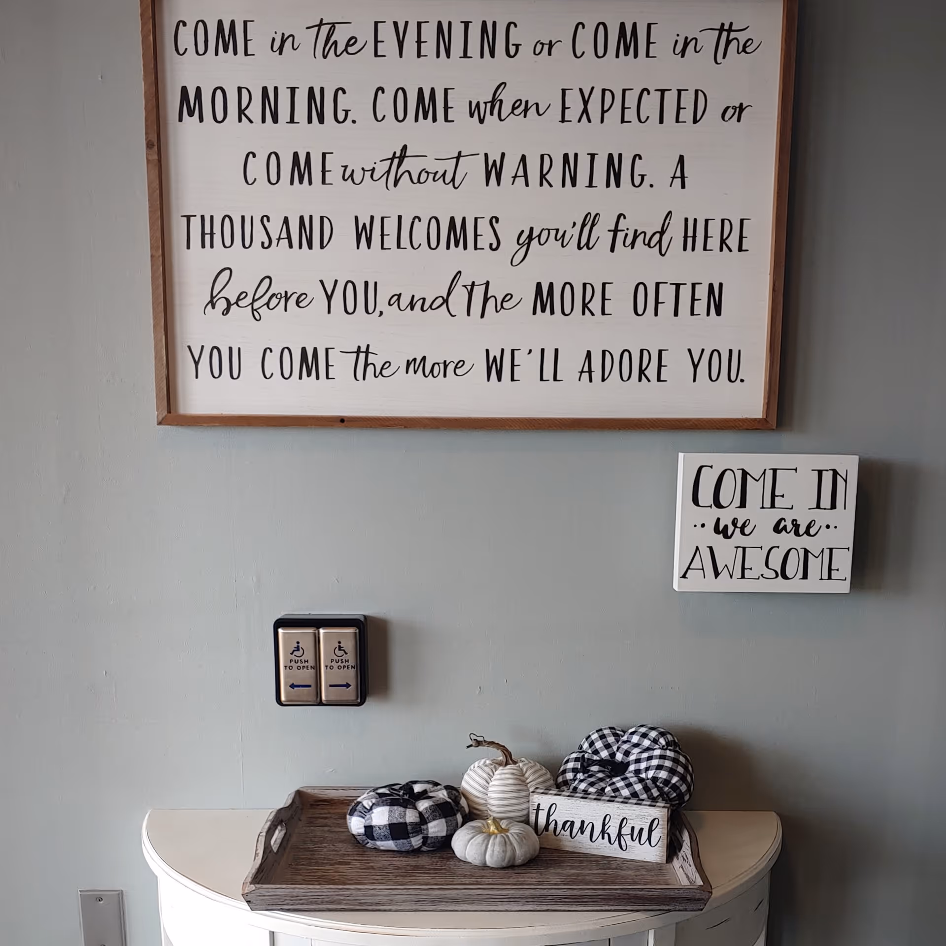 A wall with two framed signs featuring welcoming messages. The larger sign reads 'COME in the EVENING or COME in the MORNING. COME when EXPECTED or COME without WARNING. A THOUSAND WELCOMES you'll find HERE before YOU, and the MORE OFTEN YOU COME the more WE'LL ADORE YOU.' The smaller sign says 'COME IN we are AWESOME.' Below the signs is a wooden tray with decorative pumpkins and a small sign that says 'thankful.' There are also two accessibility push-to-open buttons on the wall.