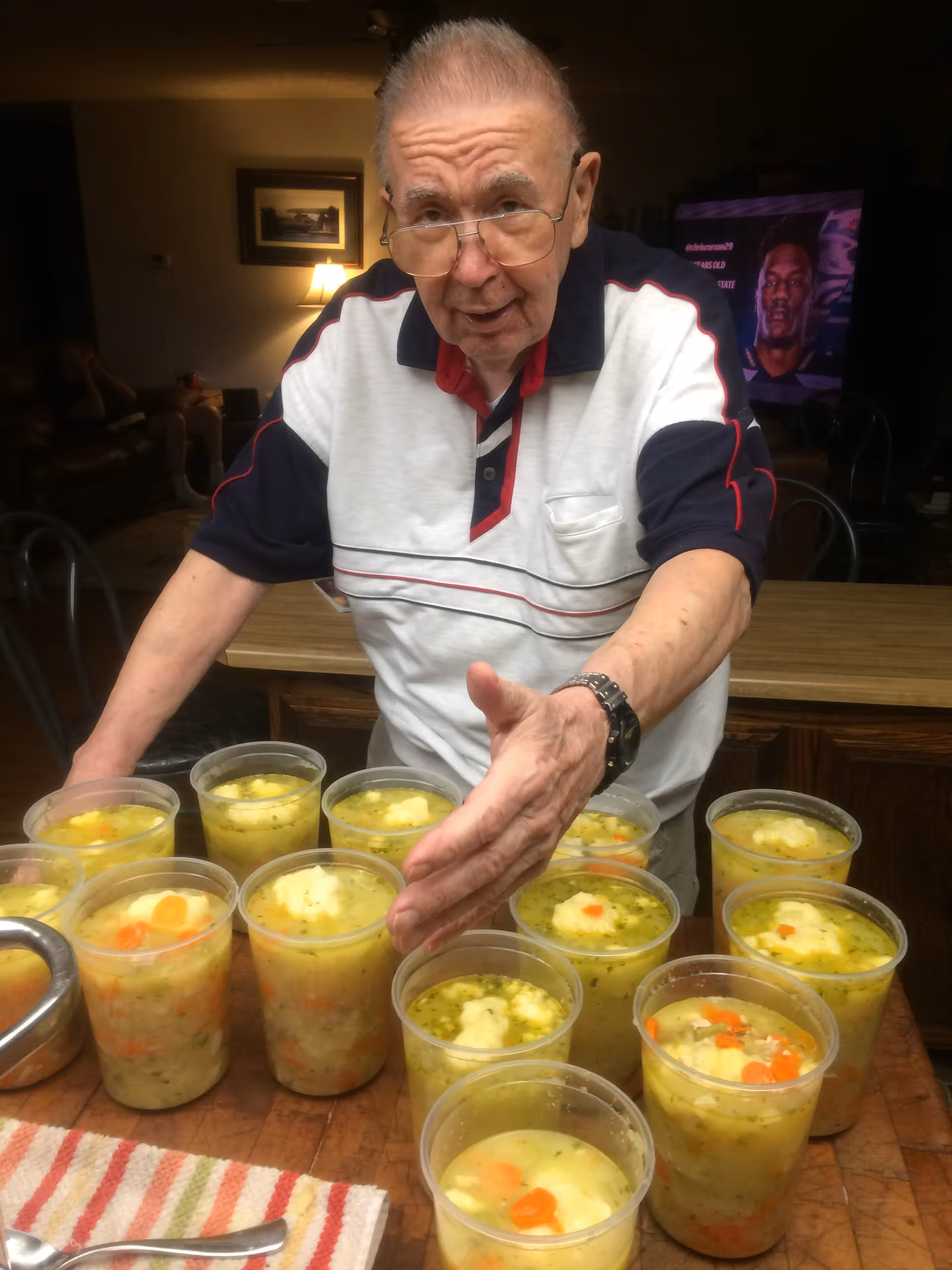 An elderly man wearing glasses and a white polo shirt with navy and red accents is standing behind a table with multiple clear plastic cups filled with soup containing vegetables and dumplings. The setting appears to be a cozy living or dining area with a lamp and framed picture in the background.