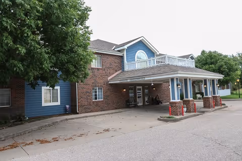 Exterior view of a two-story senior living facility building with a covered entrance supported by brick columns. The building has a combination of red brick and blue siding with white trim. There are trees and a paved driveway in front of the entrance.