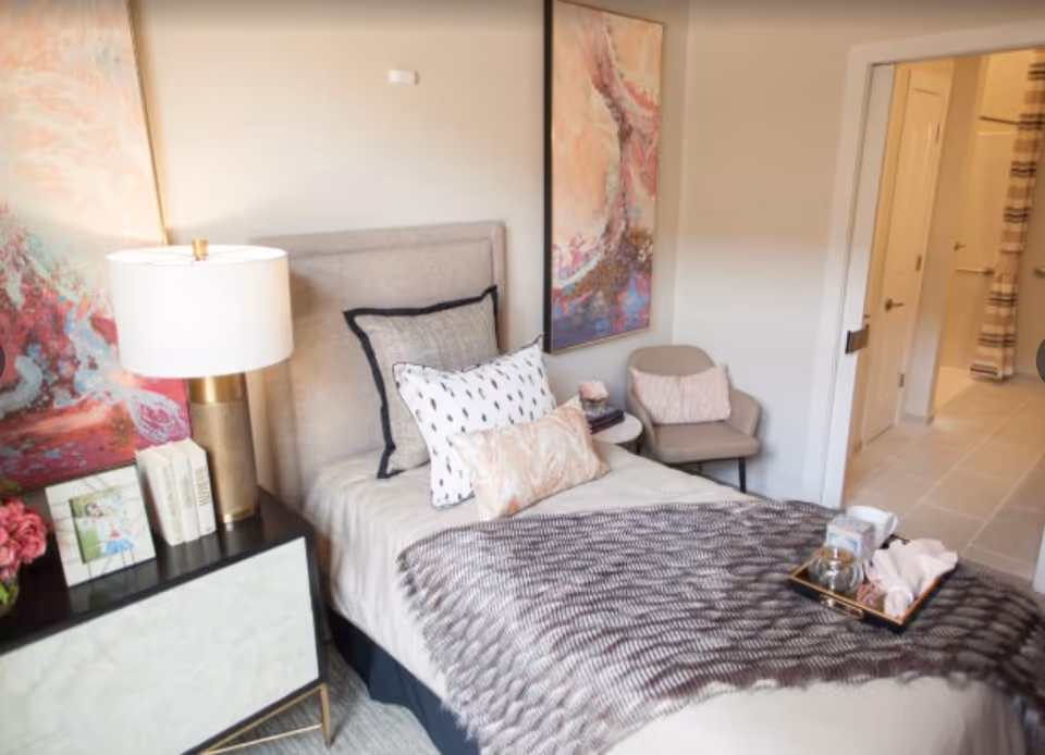 Single furnished bedroom with a made bed, bedside table and lamp, decorative artwork, and an open doorway leading to a bathroom.