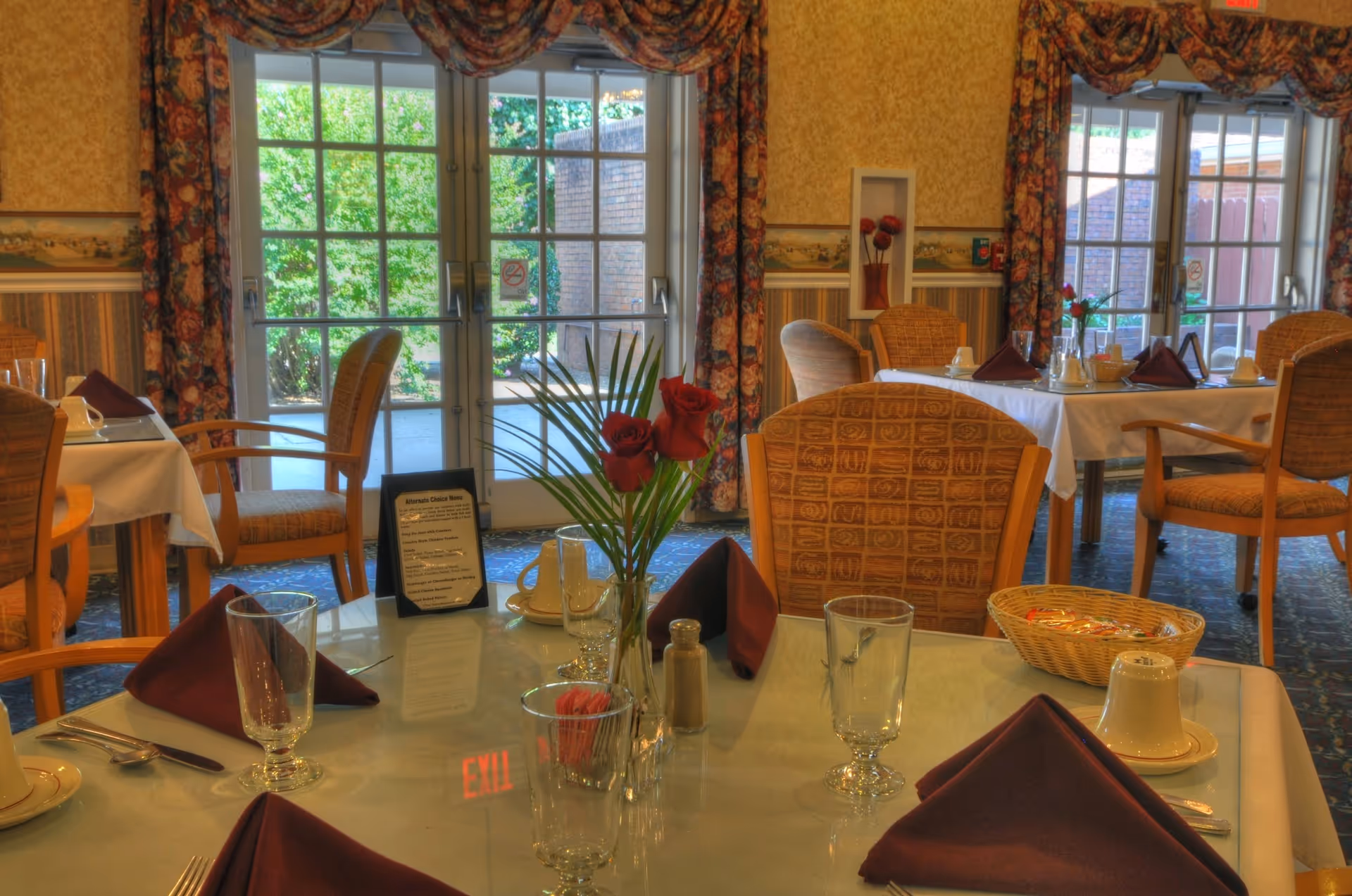 Dining room with neatly set tables, folded maroon napkins, glassware and a vase of red flowers in front of glass doors to an outdoor area.