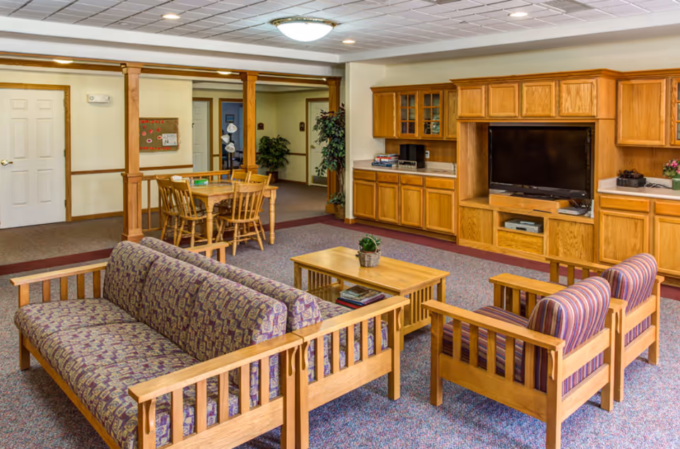 A cozy living room area in a senior living facility featuring a patterned sofa, two striped armchairs, a wooden coffee table with a small plant, and a wooden entertainment center with a flat-screen TV. In the background, there is a wooden dining table with chairs and built-in wooden cabinets along the wall.