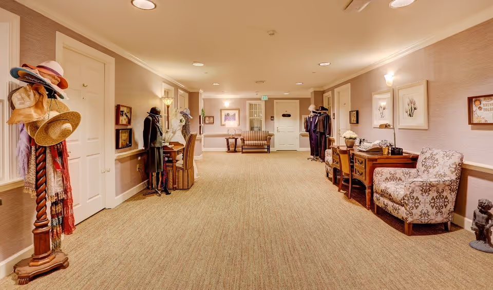 Well-lit senior living hallway with chairs, desks, and hat-and-coat stands lining a carpeted corridor.