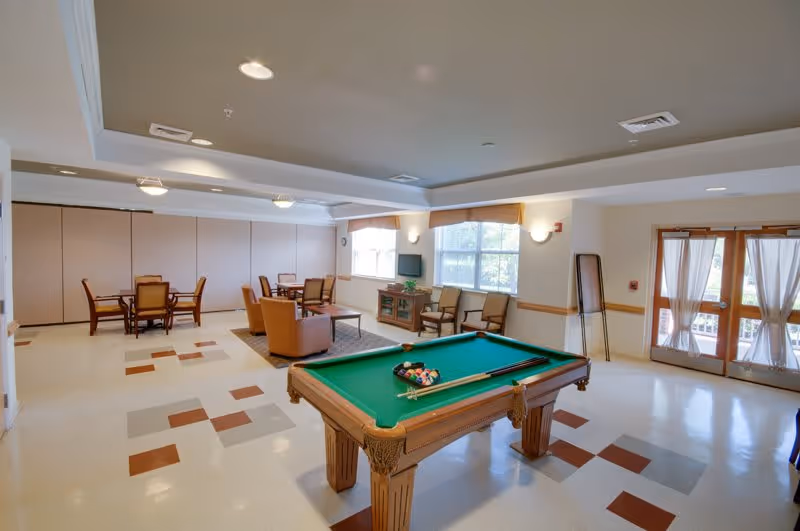 A bright common room with a pool table in the foreground and seating and dining areas near windows and doors.