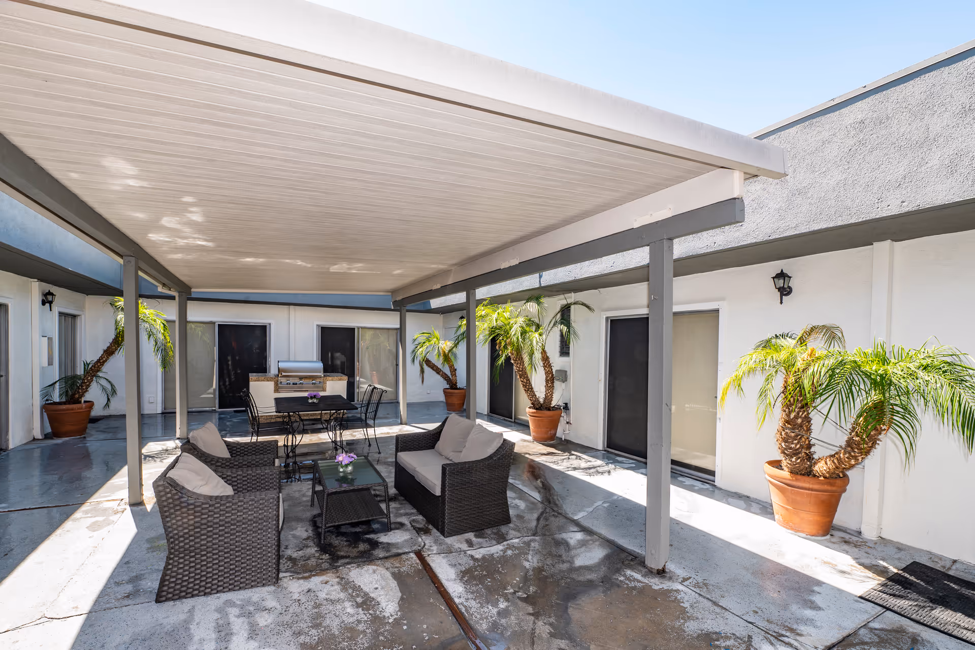 Outdoor covered patio area with wicker seating including two armchairs and a loveseat around a glass coffee table. There is a dining table with chairs and a barbecue grill in the background. Several potted palm plants line the perimeter against white walls with sliding glass doors.