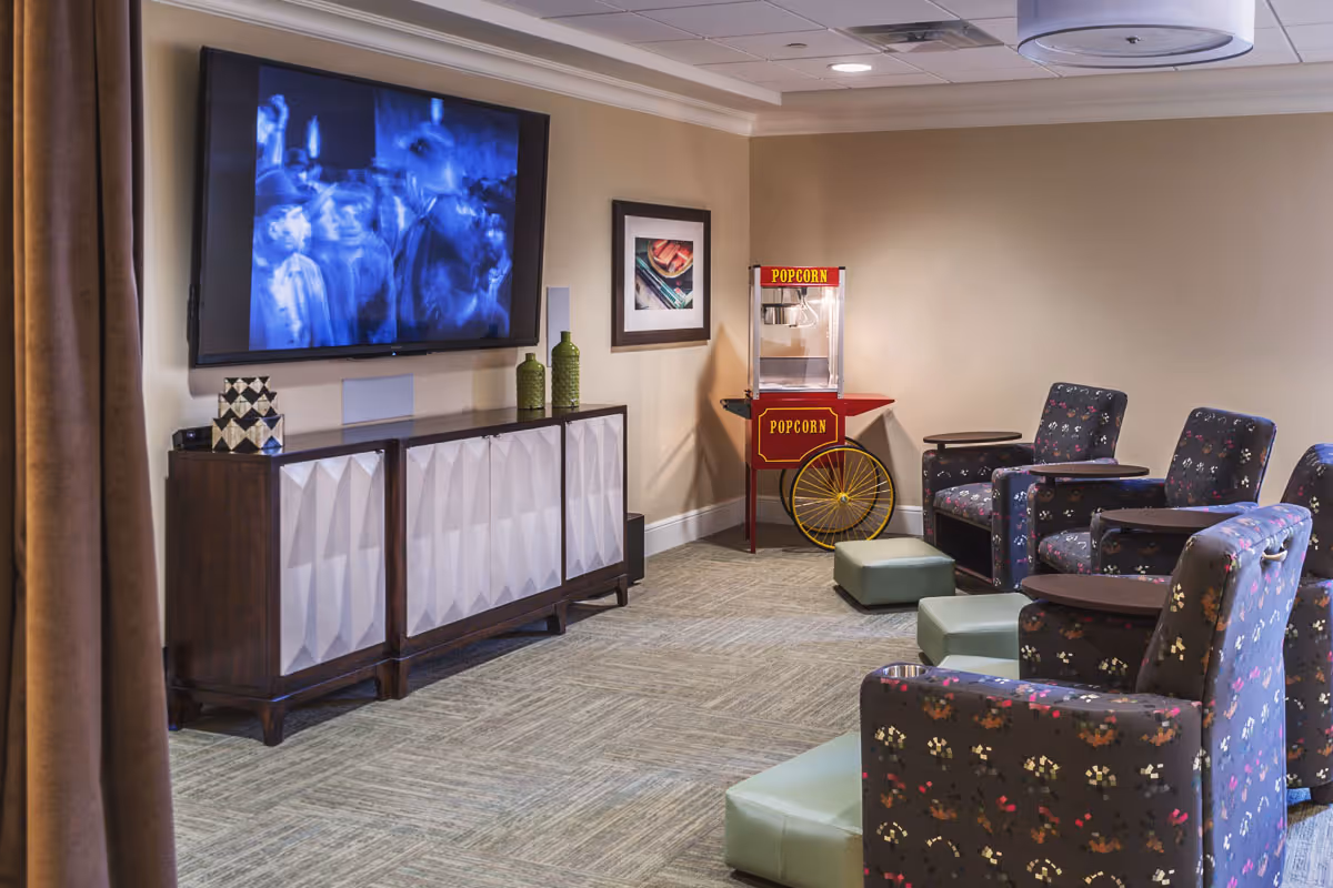 A cozy entertainment room with a large flat-screen TV mounted on the wall showing a black and white movie. Below the TV is a wooden cabinet with decorative items on top. To the right, there is a vintage-style popcorn machine and several upholstered armchairs with small attached tables, along with green ottomans on a carpeted floor.