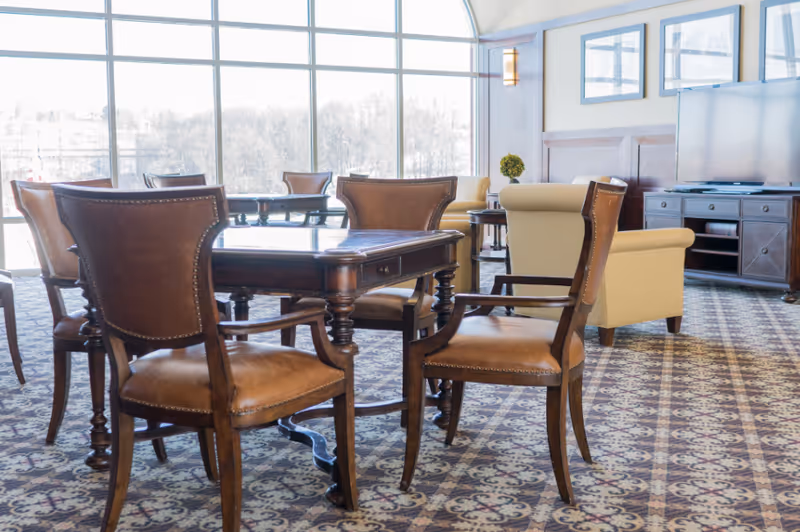Sunlit communal lounge with leather dining chairs around a wooden table, upholstered armchairs, large windows, and a TV console.