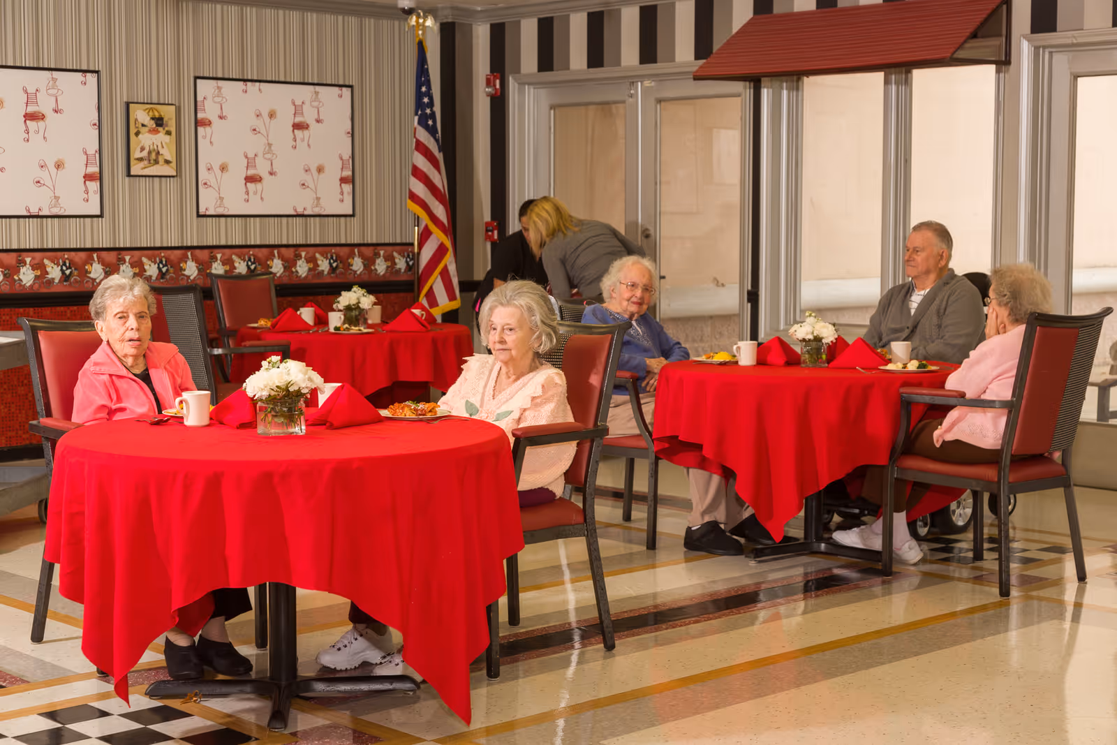 A group of elderly people sitting at two round tables covered with red tablecloths in a dining area. The tables have white floral centerpieces and red napkins. The room has striped wallpaper with framed artwork and an American flag in the background. A staff member is seen attending to something in the background.
