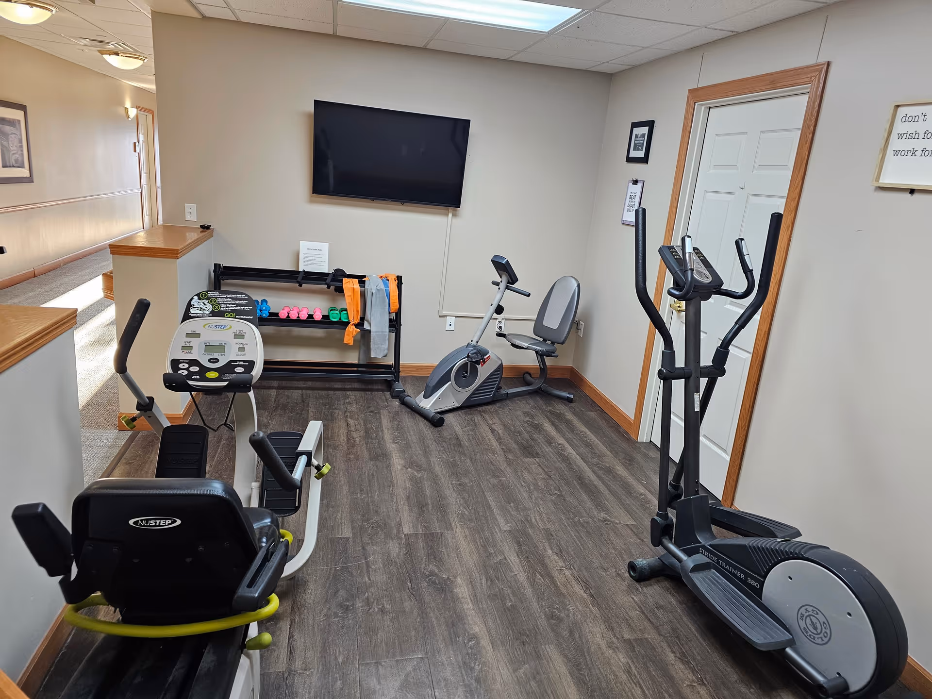 A small fitness room with exercise equipment including a recumbent bike, an elliptical machine, and a stationary bike. There is a rack with colorful dumbbells and resistance bands against the wall, and a flat-screen TV mounted above it. The room has wood flooring, beige walls, and a door on the right side.