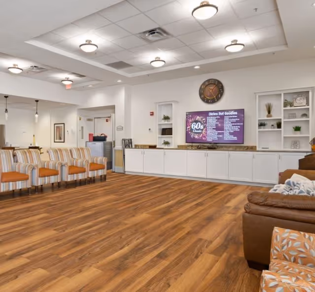 Spacious community lounge with wood flooring, rows of chairs, built-in cabinets and a wall-mounted TV beneath a clock.
