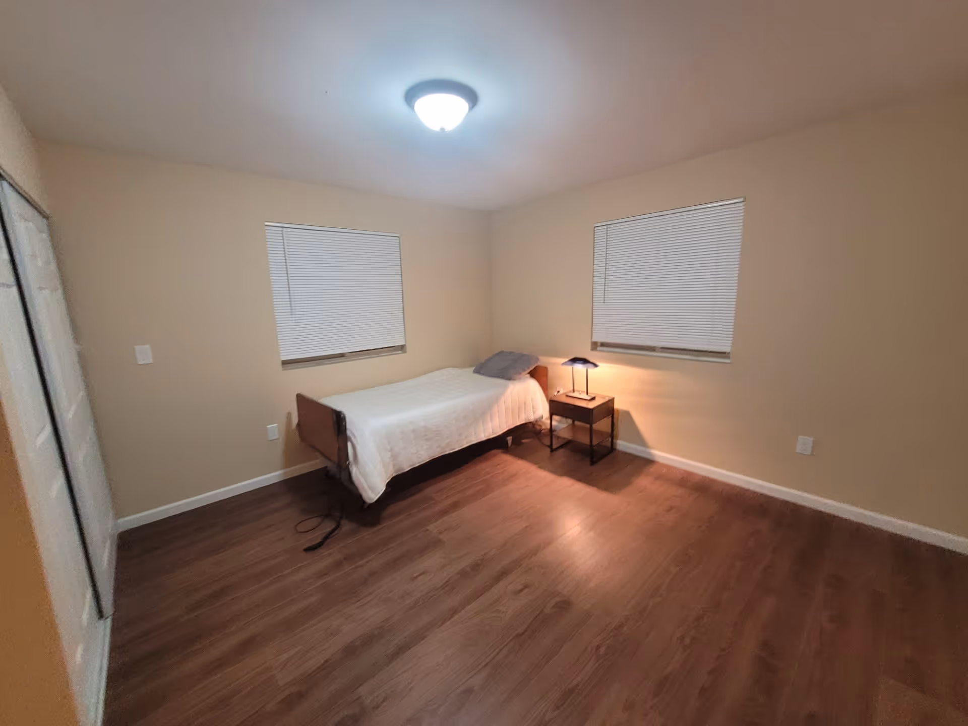 A simple bedroom with a single bed covered with a white quilt and a gray pillow. There is a small wooden nightstand with a lamp on it next to the bed. The room has two windows with closed white blinds, beige walls, and wooden flooring. A ceiling light fixture is turned on, illuminating the room.