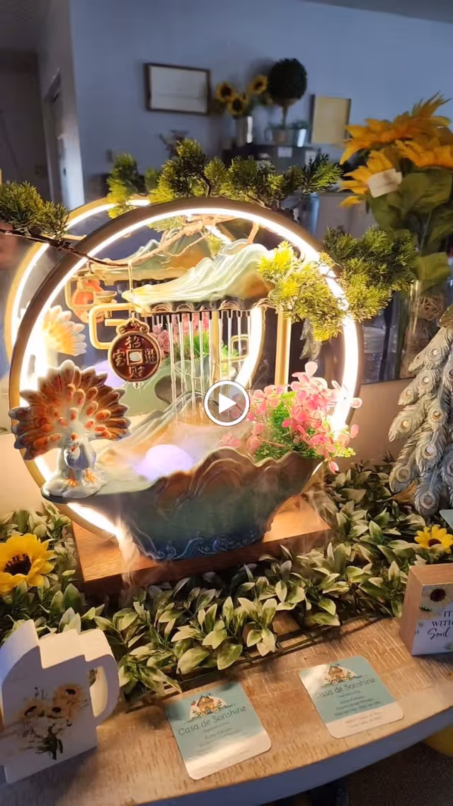 Decorative tabletop arrangement featuring a circular lighted frame with greenery, a small peacock figurine, pink flowers, and a miniature water fountain. The display is surrounded by artificial green leaves and sunflowers, with two Casa de Sonshine business cards and a small white decorative watering can with sunflower design on the wooden table. The background shows a softly lit room with more sunflowers and framed pictures on the wall.