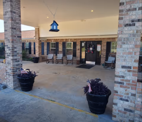 Covered entrance area of a brick building with several rocking chairs lined up against the wall, two large black planters with purple plants and small American flags, and a hanging blue bird feeder. The entrance door is decorated with a wreath.