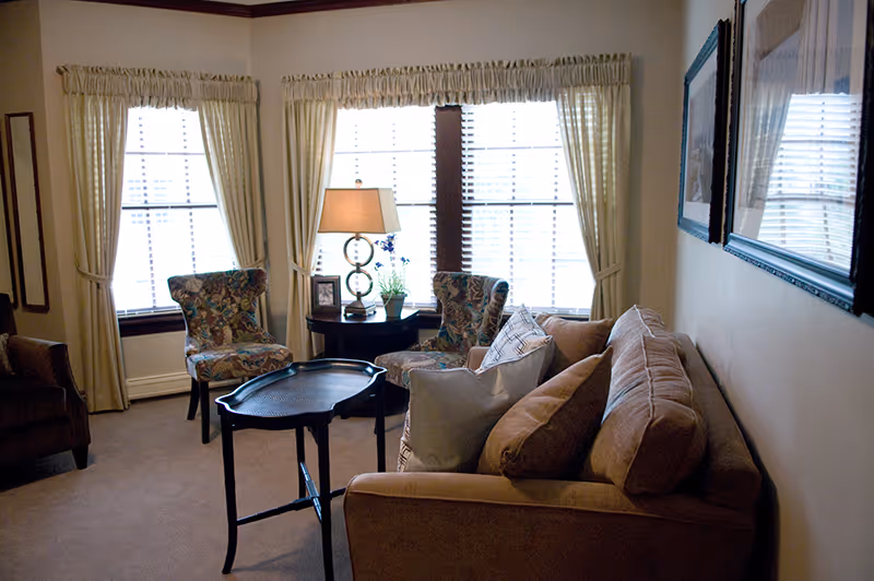 Cozy living room with a sofa, patterned armchairs, a side table and lamp in front of three large curtained windows.