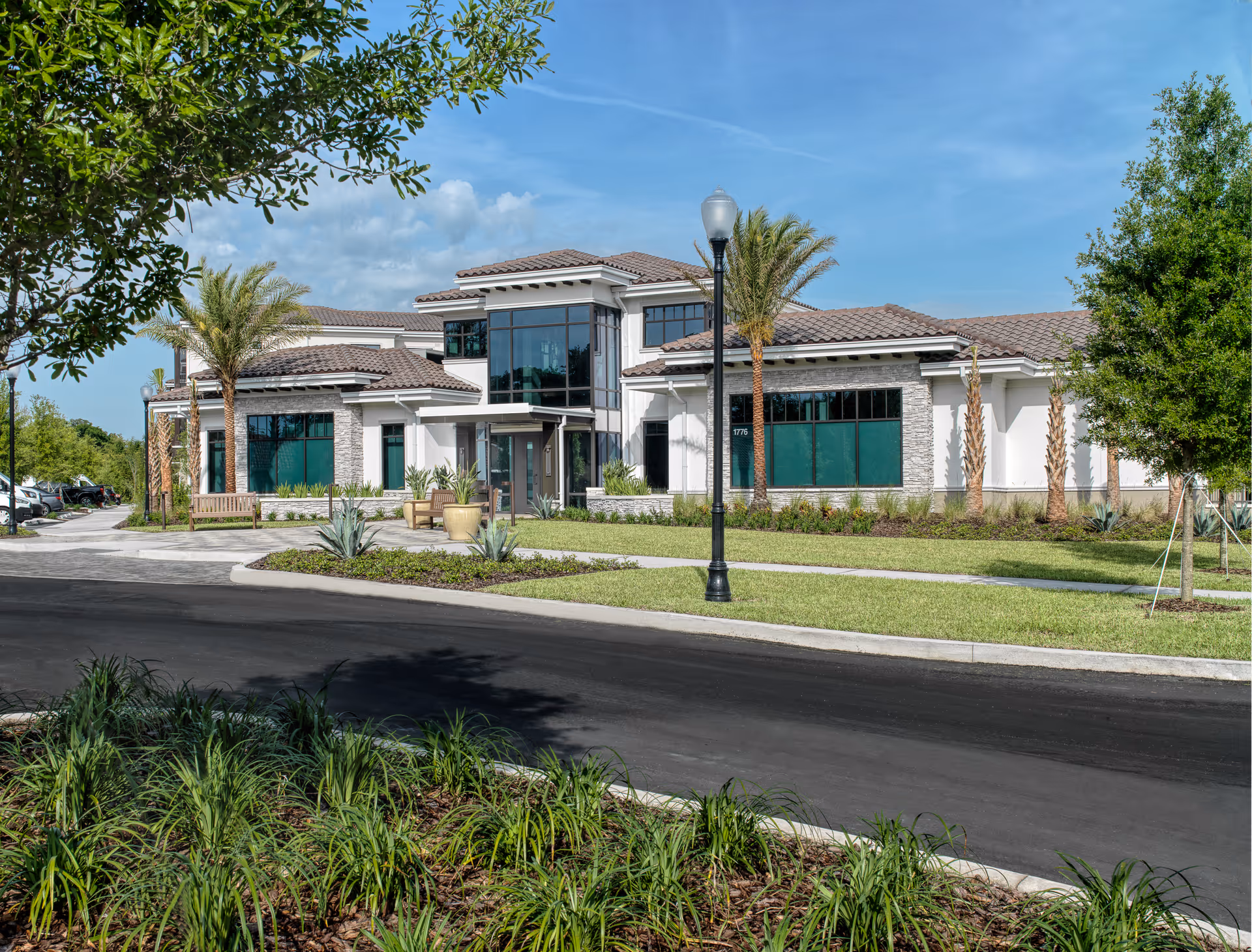 Front exterior of a modern senior living facility building with palm trees, lawn, benches, and a driveway.