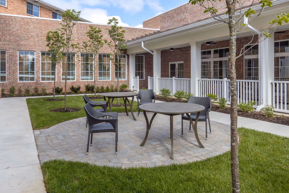 Outdoor courtyard area at Sayre Christian Village featuring two round tables with four black wicker chairs on a circular paved patio surrounded by green grass, young trees, and a brick building with large windows and white railings in the background under a partly cloudy sky.