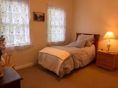 A cozy bedroom with a single bed covered in beige bedding and a white blanket at the foot. There are two windows with sheer white curtains letting in natural light. A wooden nightstand with a lamp and a book is beside the bed. A small wooden chair and a dresser with a flower vase are also visible.