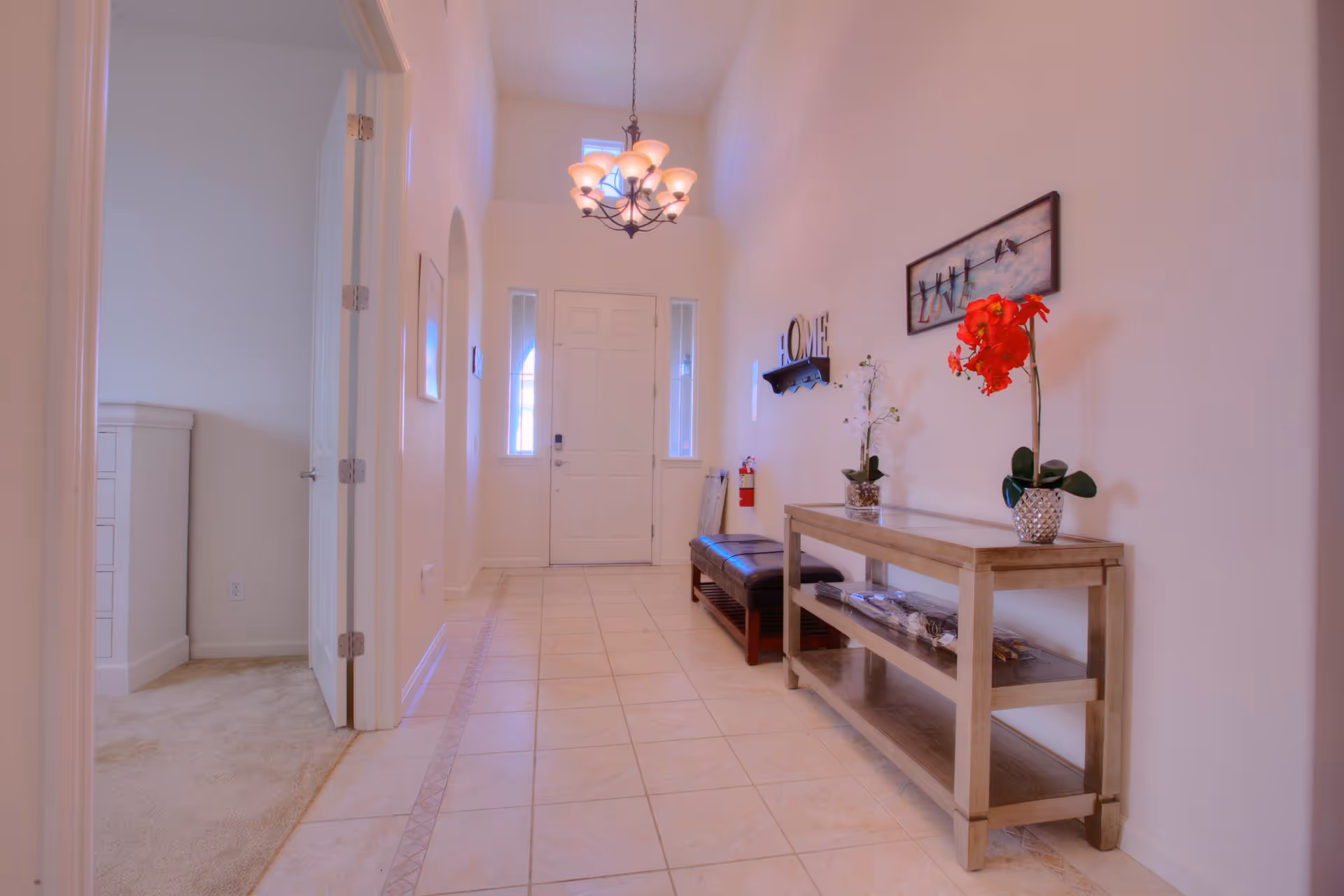 A bright hallway with tiled floor leading to a white front door with glass panels on each side. On the right side, there is a wooden console table with two potted flowers and a bench underneath. Wall decorations include a 'HOME' sign and a framed picture with the word 'LOVE'. On the left, an open door reveals a carpeted room with white furniture.