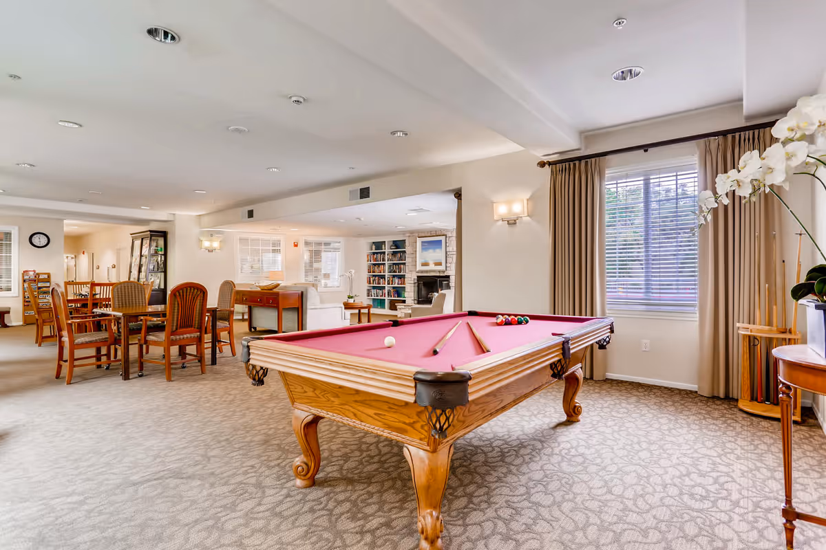 A spacious common area in a senior living facility featuring a pool table with a red felt top in the foreground. Behind the pool table, there are several wooden chairs and tables, a bookshelf filled with books, a fireplace, and large windows with beige curtains allowing natural light to enter. The room has a carpeted floor and soft ceiling lighting.