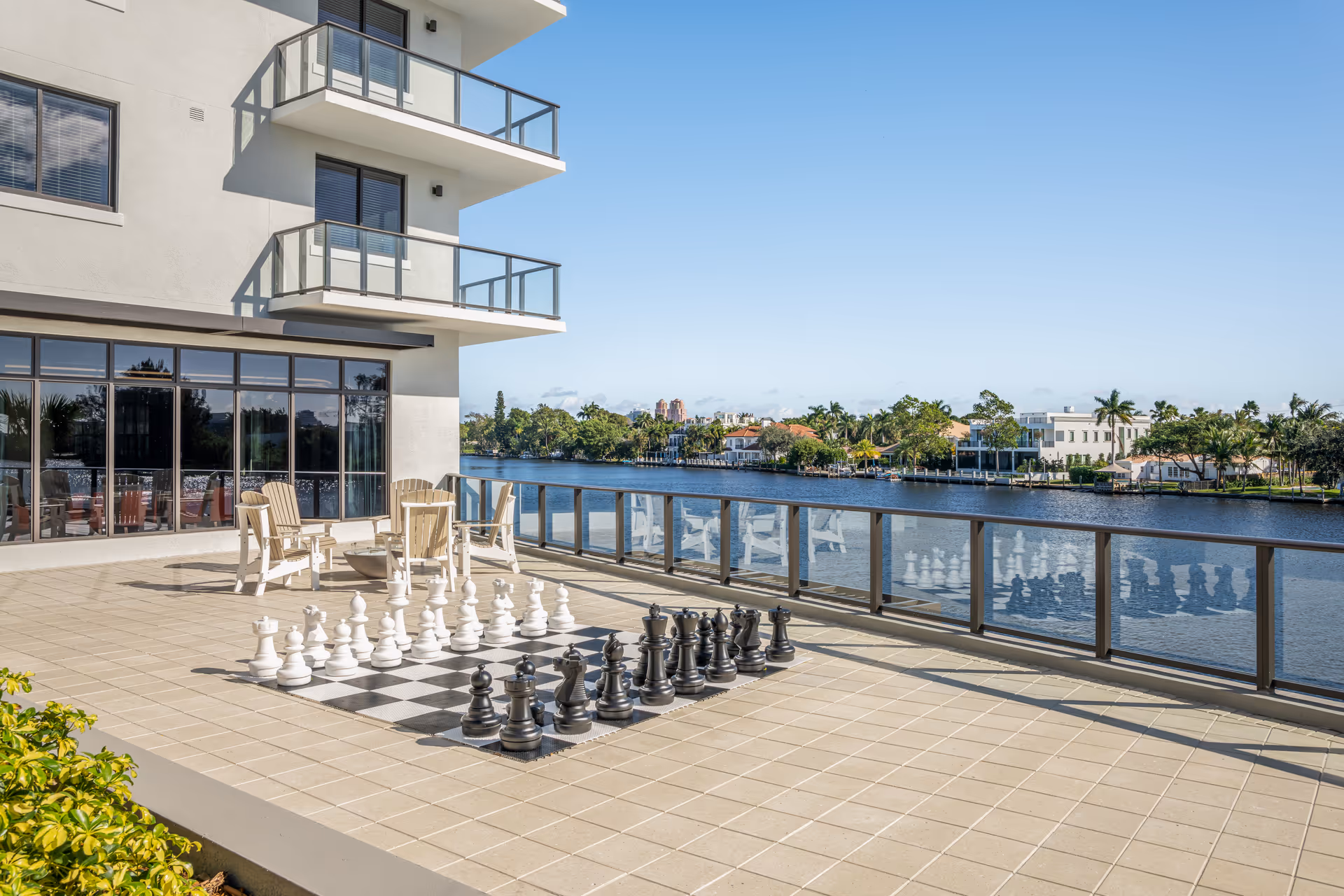 Outdoor patio area at Allegro – Fort Lauderdale, FL with a large chessboard and oversized chess pieces on the tiled floor. Several Adirondack chairs are arranged near the building with glass balconies above. The patio overlooks a calm waterway with houses and palm trees on the opposite shore under a clear blue sky.