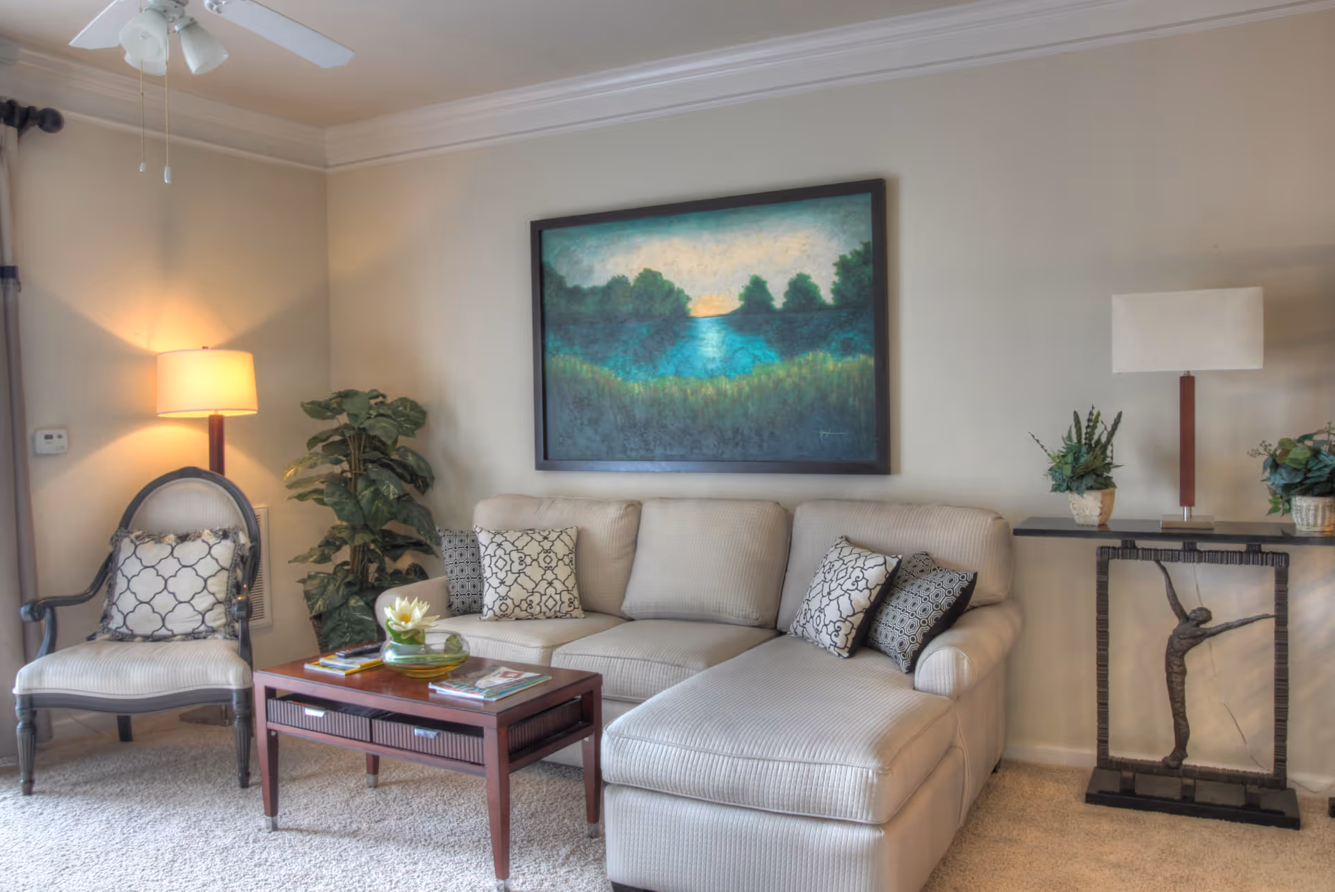 A cozy living room with a beige sectional sofa adorned with patterned throw pillows, a wooden coffee table with magazines and a decorative bowl, a cushioned armchair with a patterned pillow, a floor lamp, a potted plant, a framed landscape painting on the wall, and a side table with a lamp and two small potted plants.