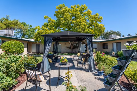 Sunlit courtyard featuring a central gazebo with patio chairs and a small table, surrounded by gardens and a single-story building.