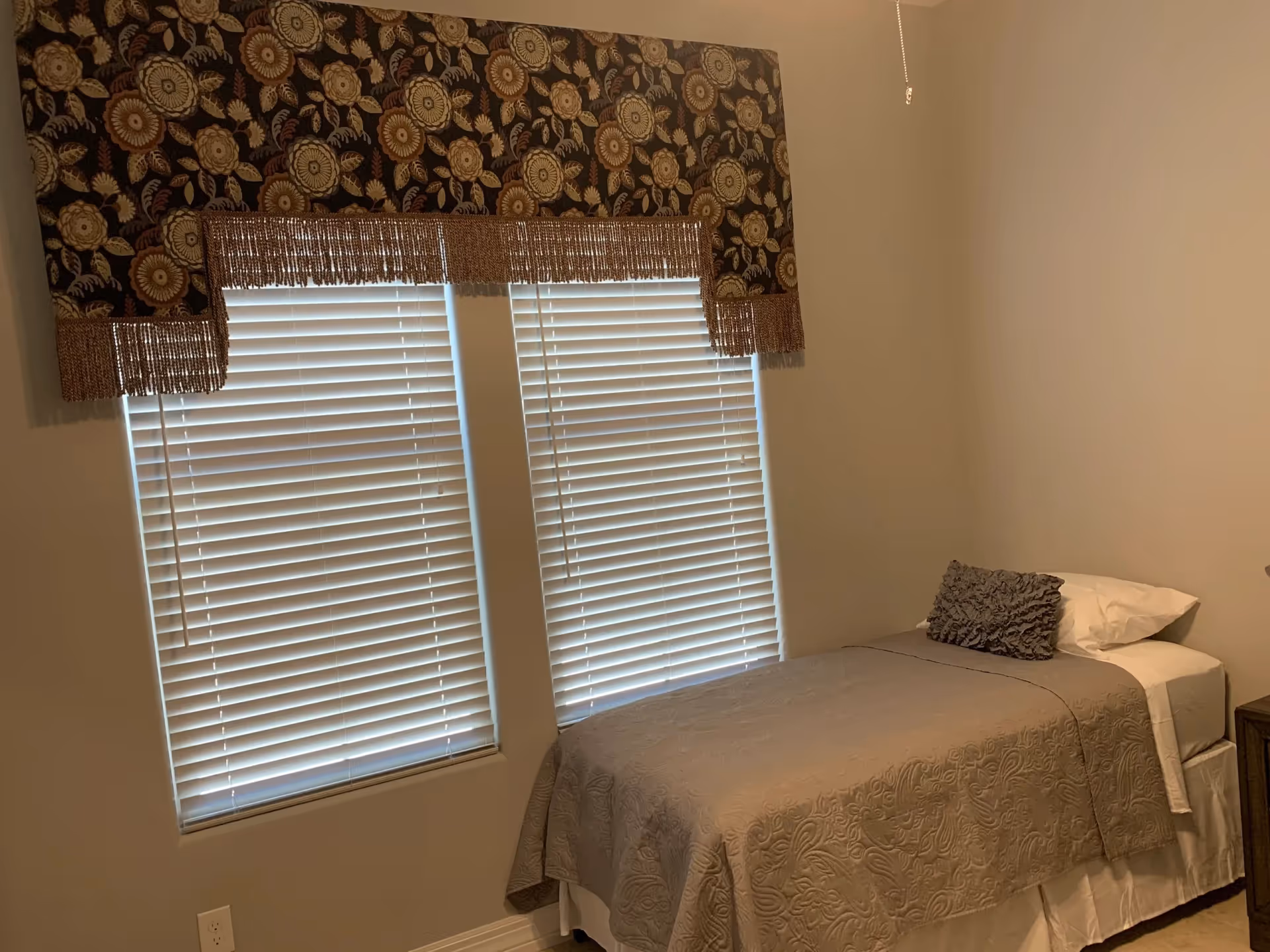 A small bedroom with two windows, a floral valance, blinds, and a bed