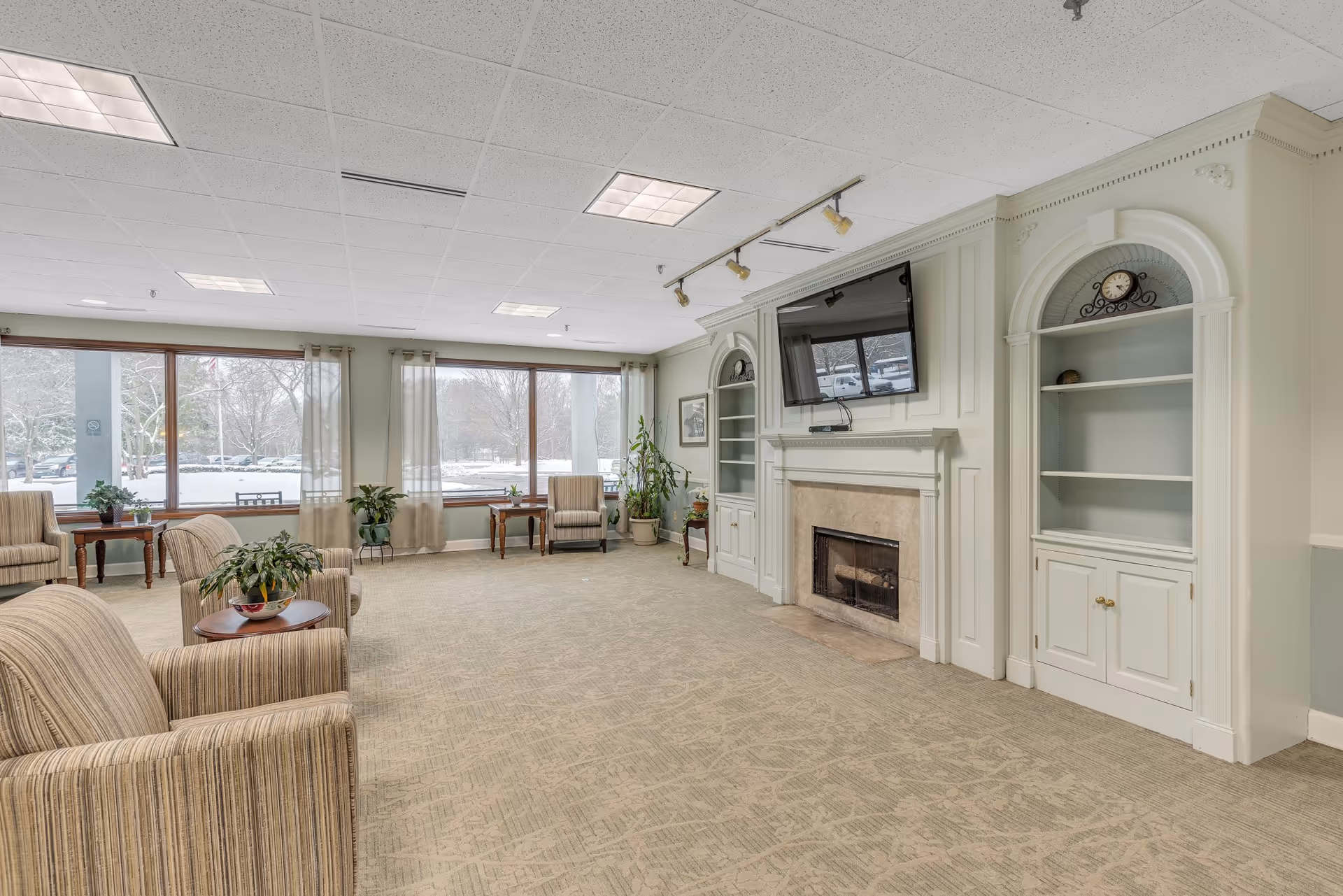 Bright communal sitting room with striped armchairs, side tables, large windows overlooking snow, a fireplace with built-in shelves and a wall-mounted TV.