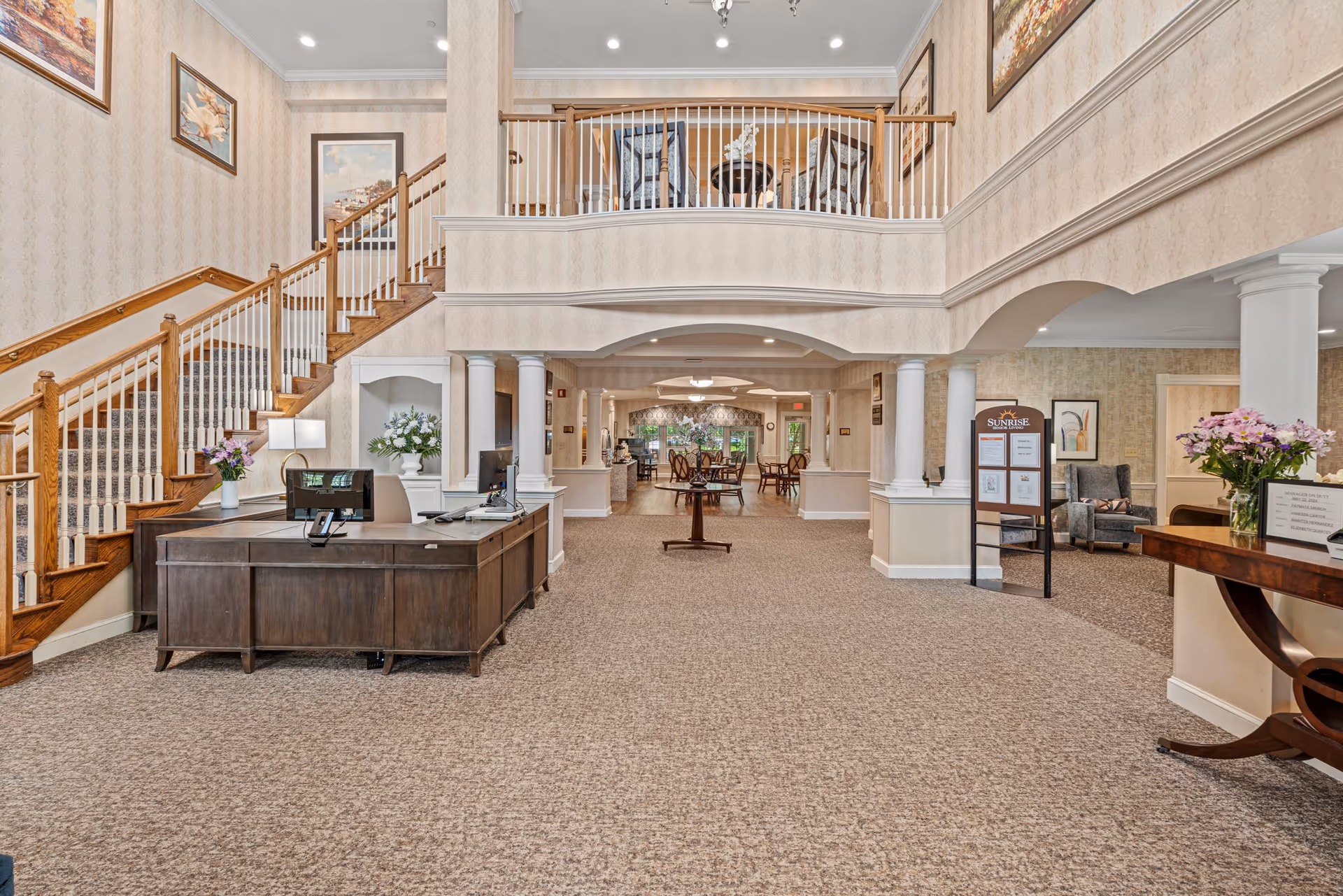Spacious two-story senior living lobby with reception desks, a grand staircase, columns, seating areas, and floral arrangements.