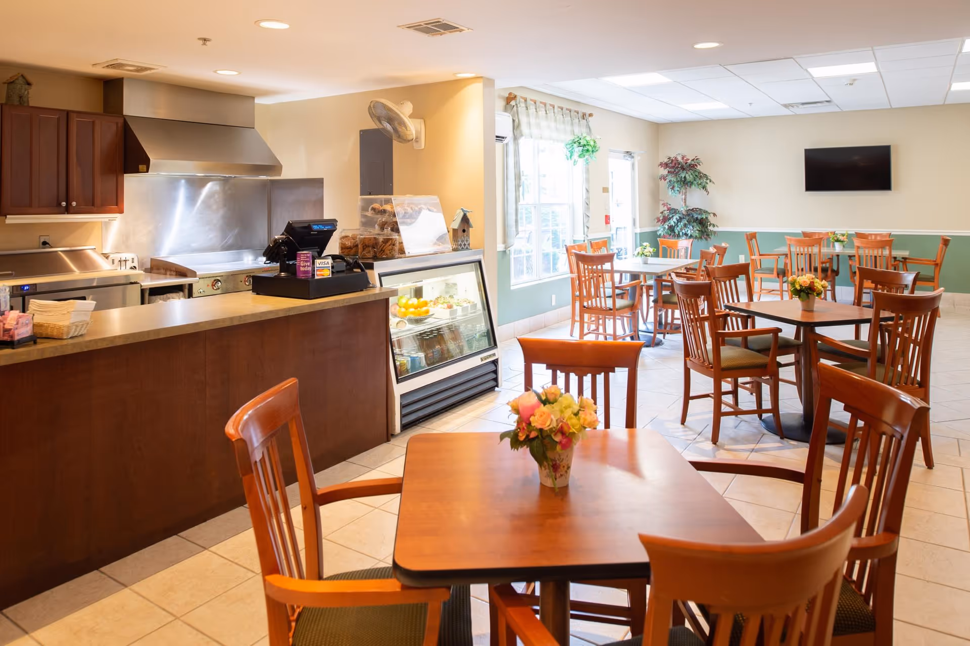 A bright dining area with wooden tables and chairs, each table decorated with a small floral arrangement. To the left, there is a kitchen counter with a cash register, a display case with baked goods, and a refrigerated display with fruits and drinks. The room has large windows letting in natural light, a wall-mounted TV, and a potted plant in the corner.