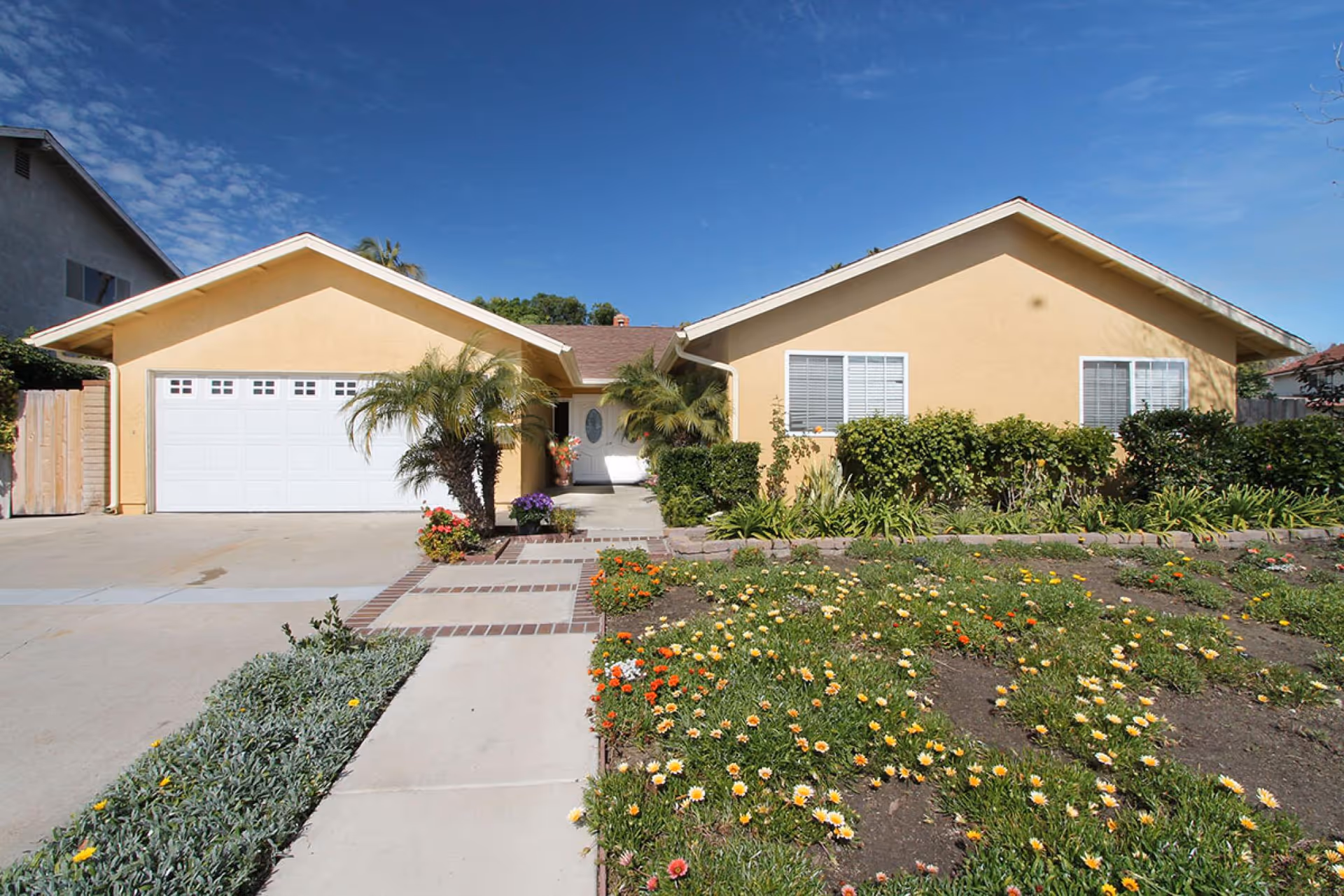 Front exterior view of a single-story beige house with a white garage door, a concrete driveway, a walkway lined with bricks, and a garden with flowering plants and shrubs under a clear blue sky.