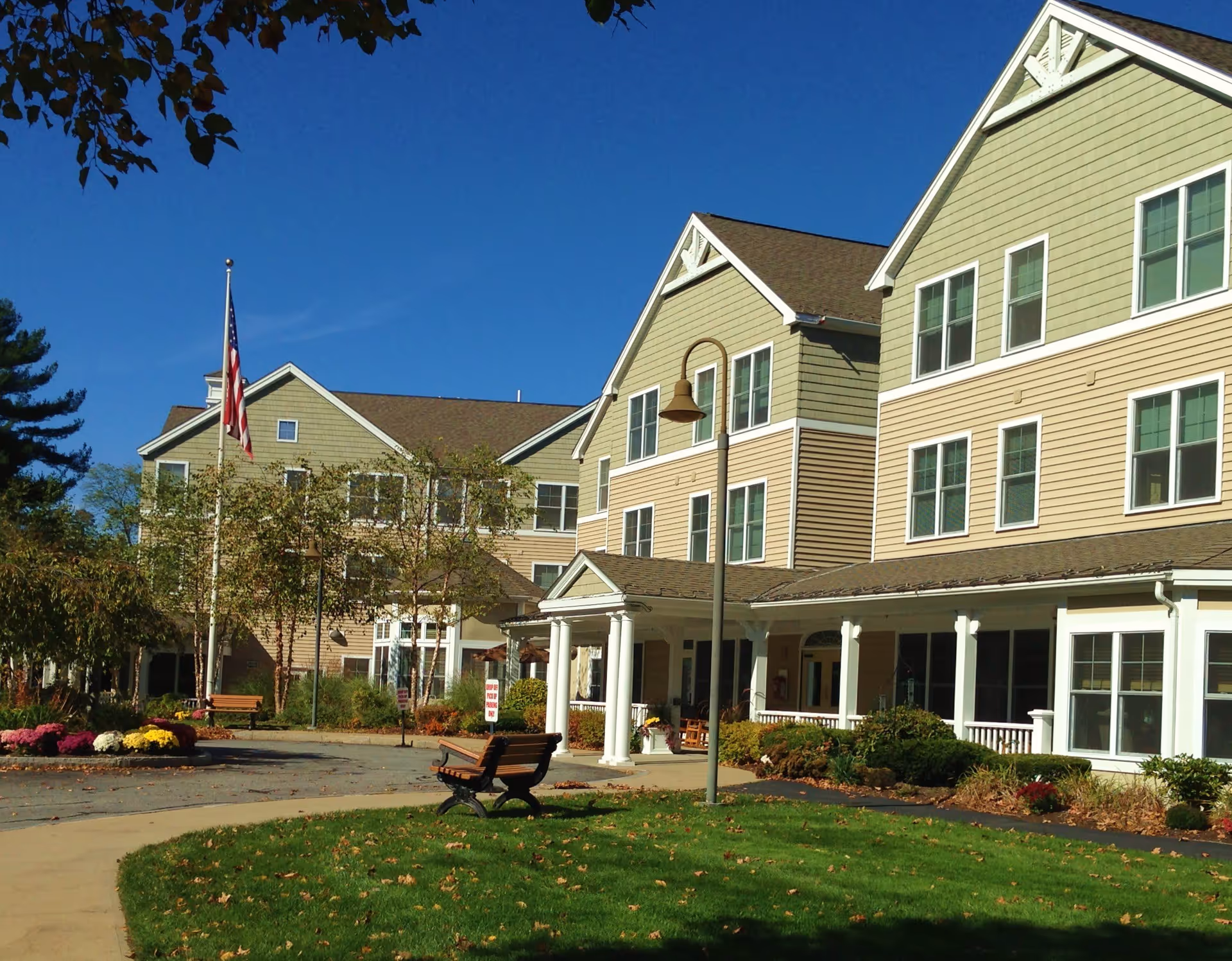 Exterior view of a senior living facility with beige siding and white trim, featuring multiple windows and a covered entrance supported by white columns. There is a flagpole with an American flag, benches, a streetlamp, and landscaped greenery including bushes and flowers under a clear blue sky.